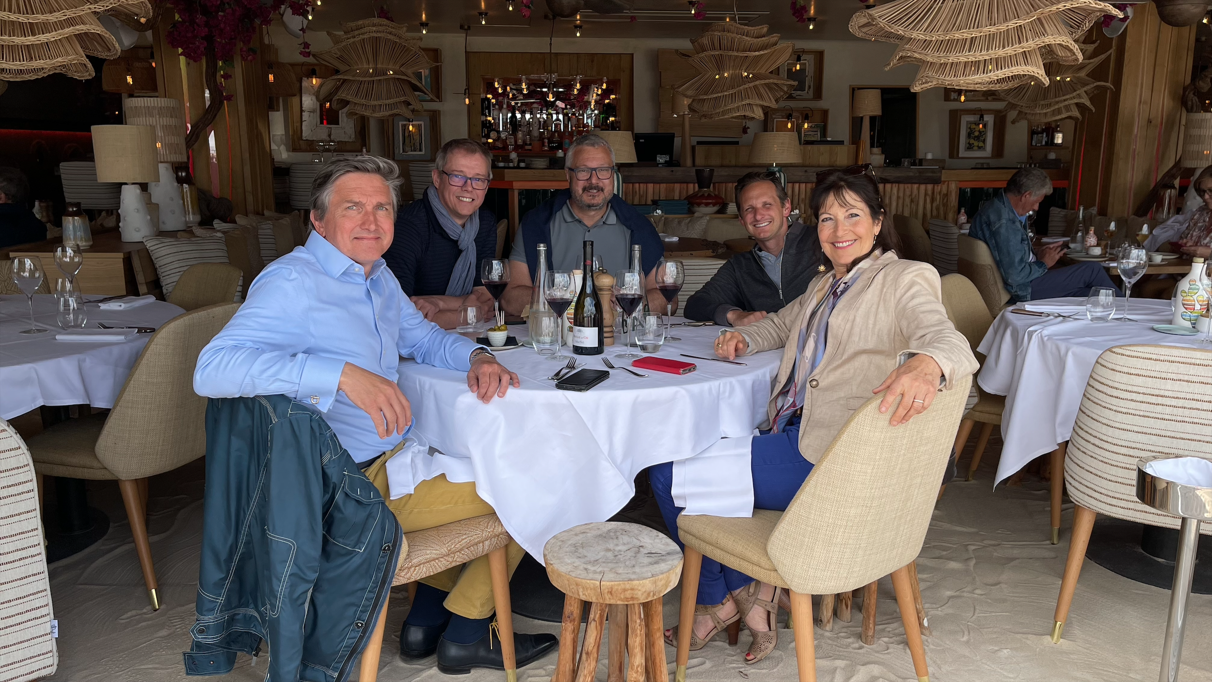 Fünf Menschen sitzen an einem runden Esstisch in einem eleganten Restaurant, mit Wein, Gläsern und Besteck, während im Hintergrund weitere Gäste dinieren.