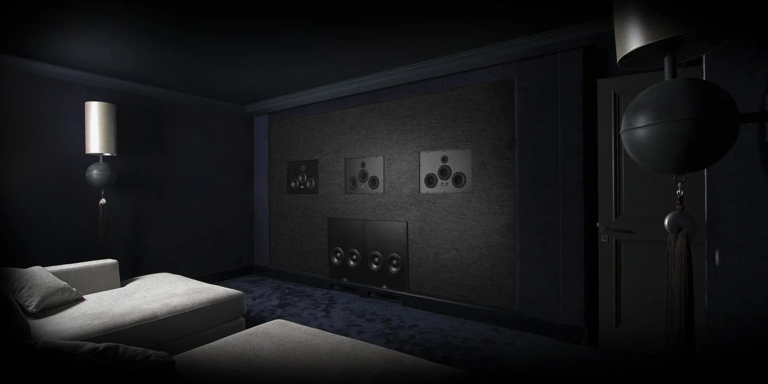 Dunkler Heimkino-Raum mit grauer Couch, Wandbilder mit Lautsprechern, Stehlampe, Deckenlampe und dunklen Wänden.