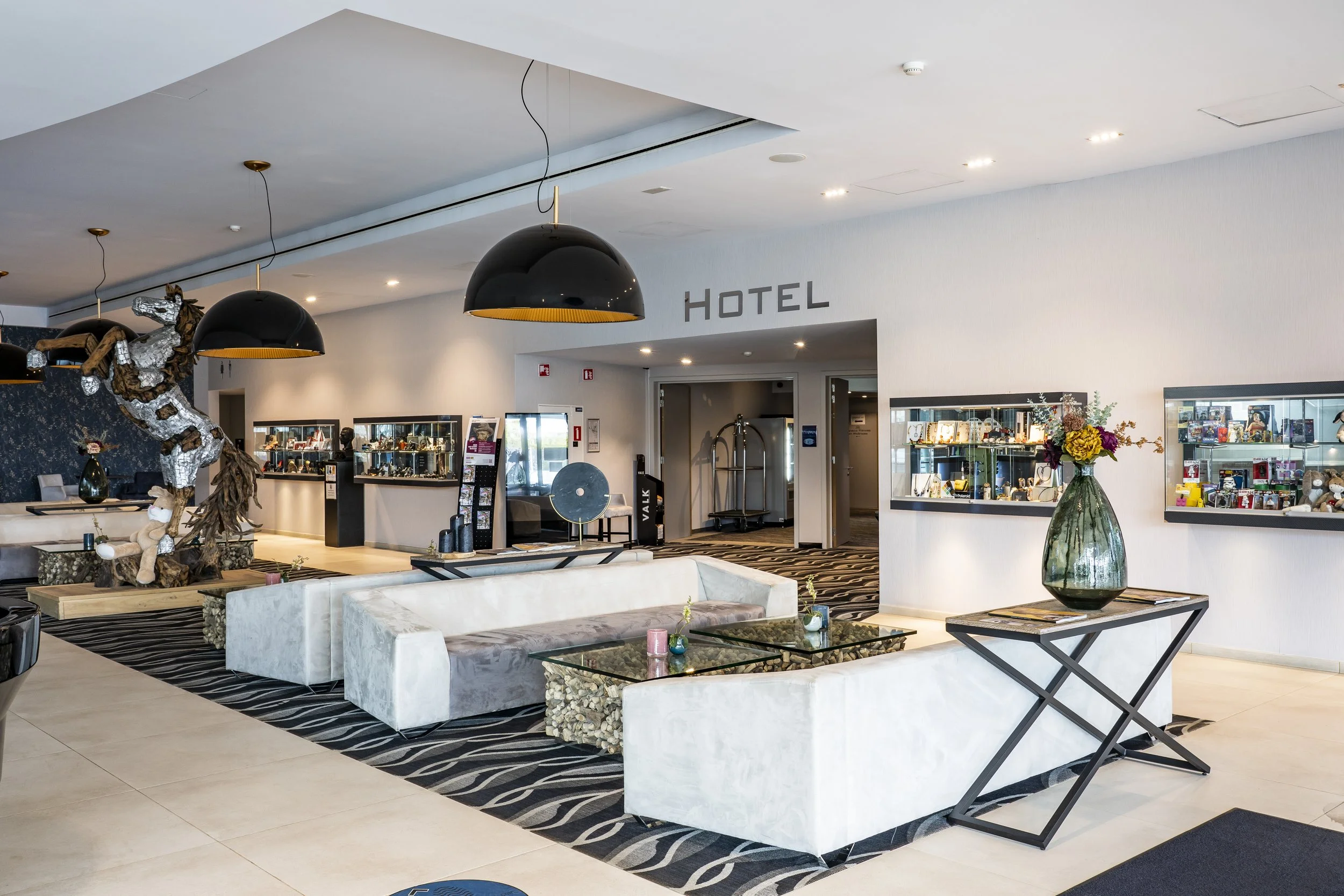 Hotel Lobby mit weißen Sofas, Couchtisch, Vampirstatue, dunkle Deckenlampen, Vitrine mit Geschenken, Blumen auf kleinem Tisch, Hotellogo an der Wand.
