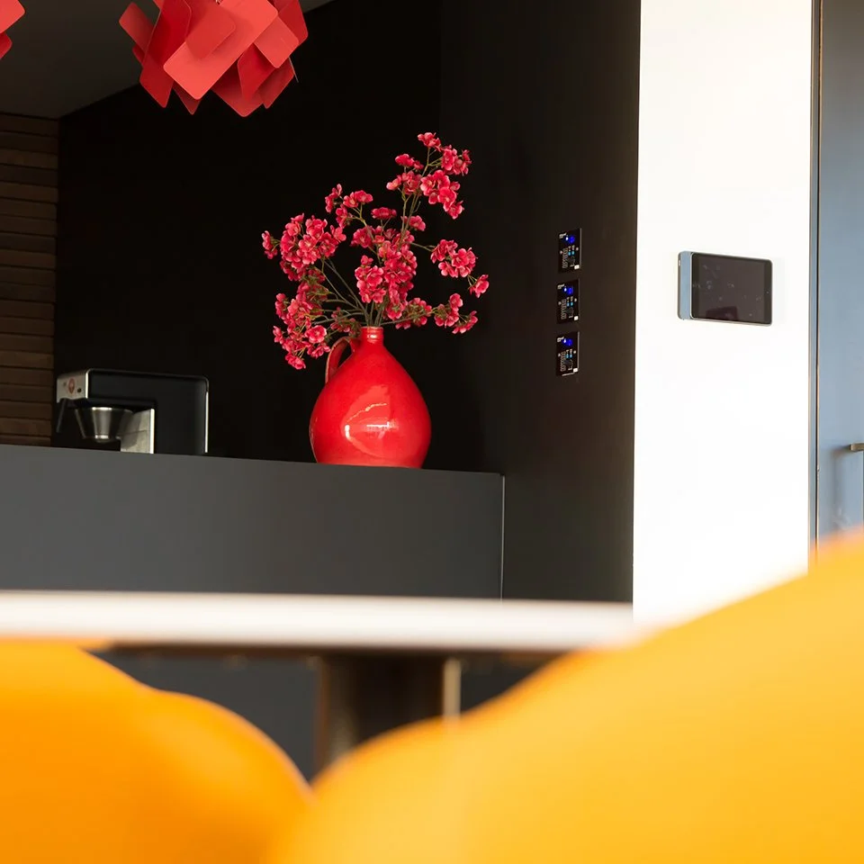 Rote Vase mit pinken Blüten auf einer schwarzen Theke, dunkle Wand, leere Tasse, digitaler Steuerungsknopf, Smartphones an der Wand, Teil eines gelben Stuhls im Vordergrund, Deckenleuchte