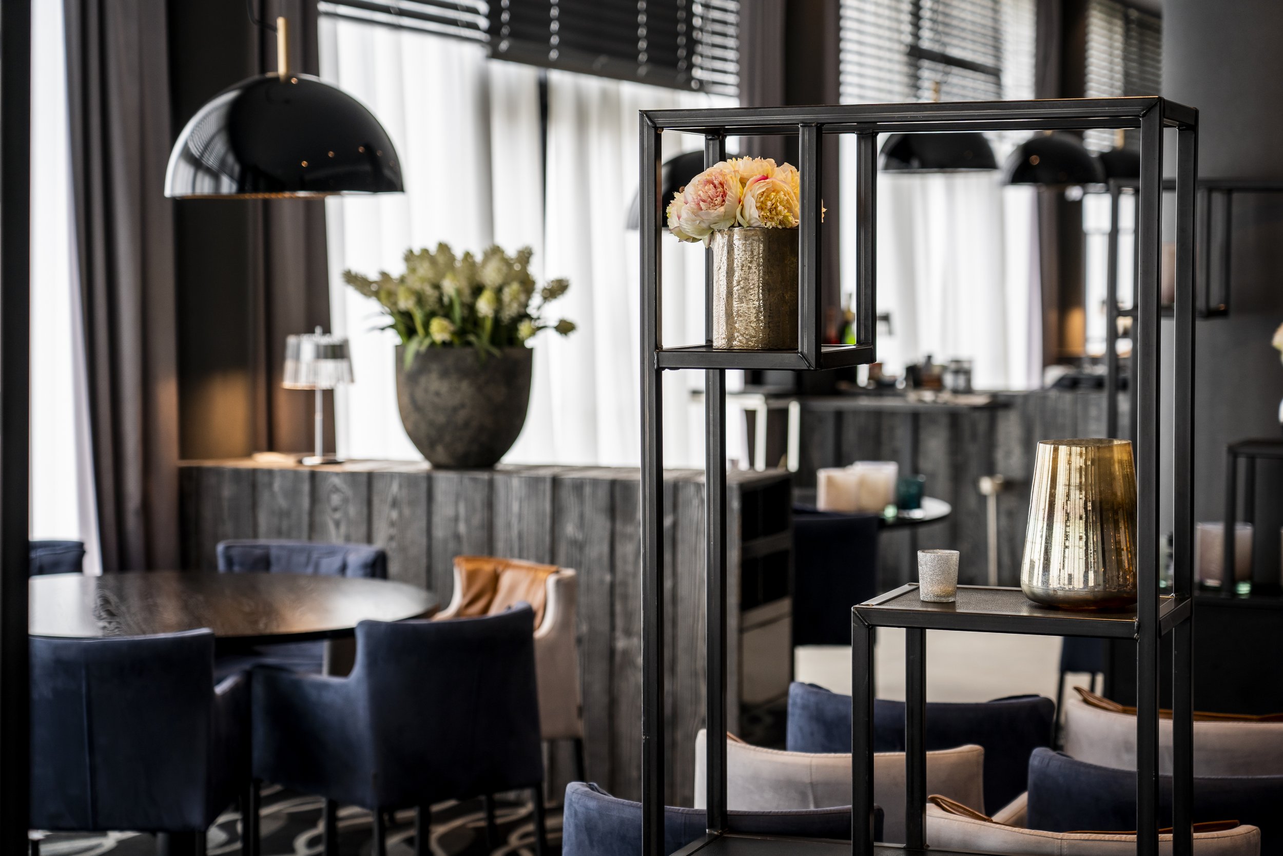 Innenraum eines stilvollen, modernen Restaurants mit dunklen Wänden, dunklen Vorhängen, gedecktem Tisch, bunten Stühlen und dekorativen Vasen mit Blumen auf schwarzem Metallregal.
