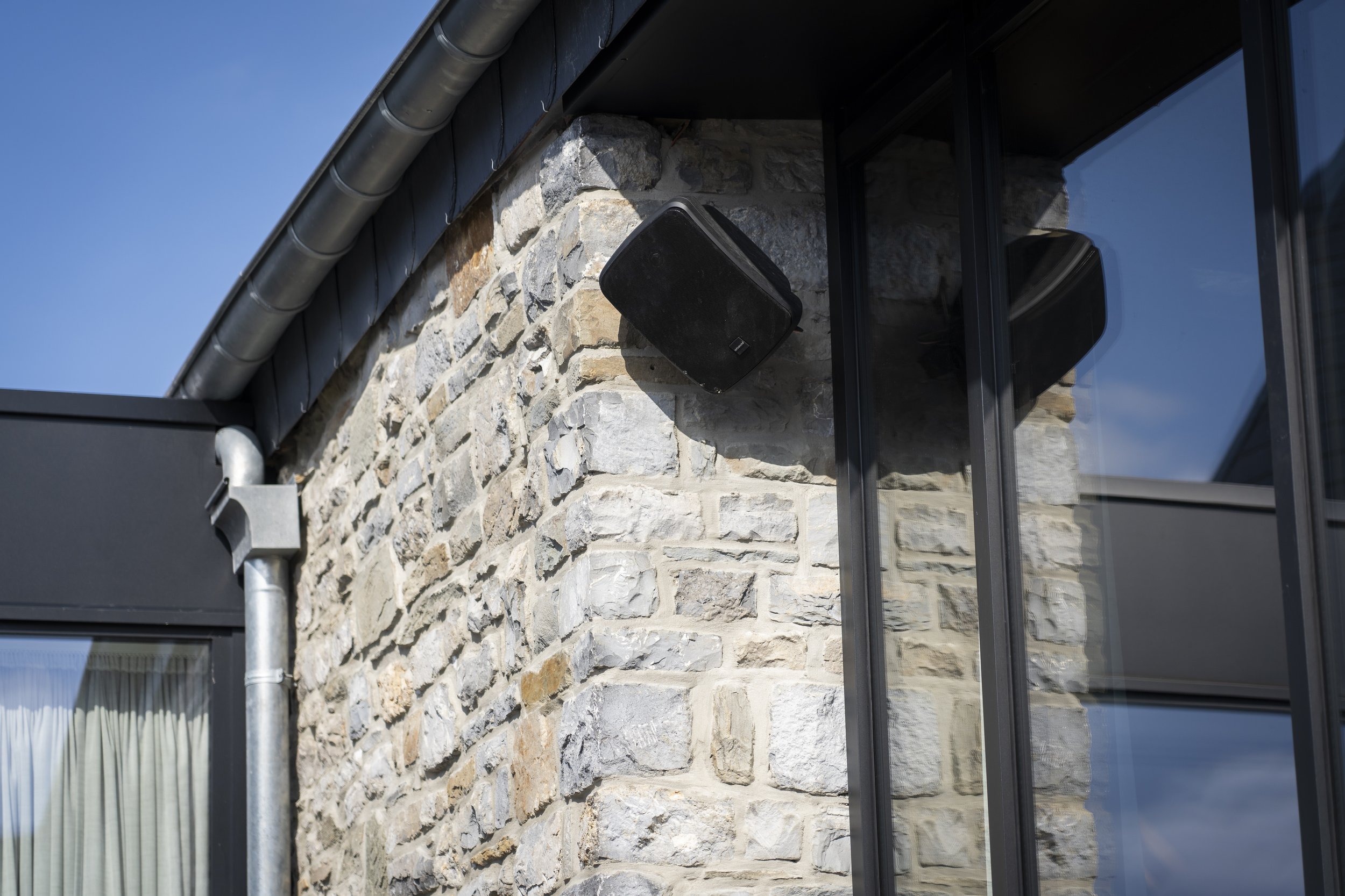 Ein Haus mit Steinwand, schwarzem Lautsprecher und großen Glasfenstern, Dach mit Dachrinnen