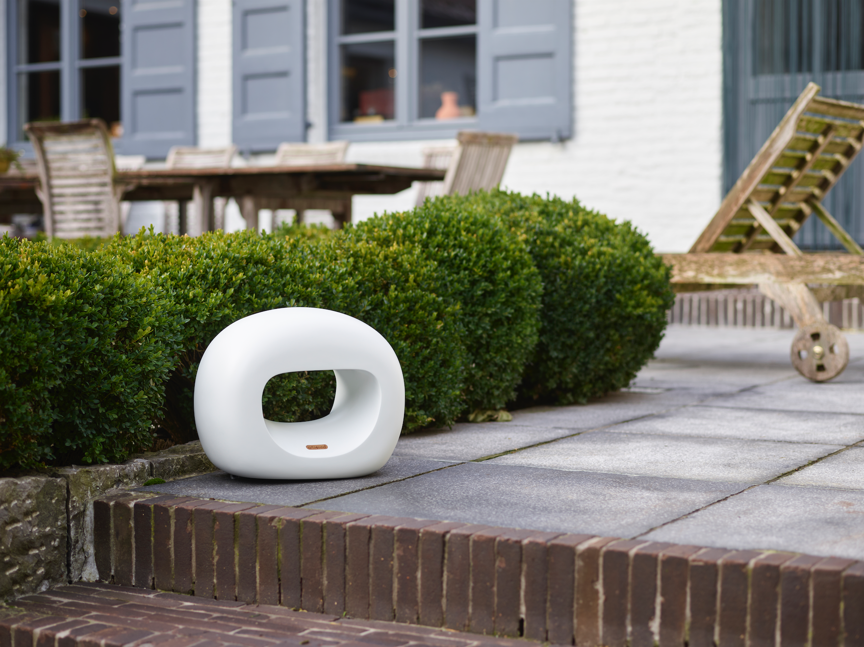 Ein weißes, modernes Gerät steht auf Pflastersteinen neben einem grünen Busch im Garten.