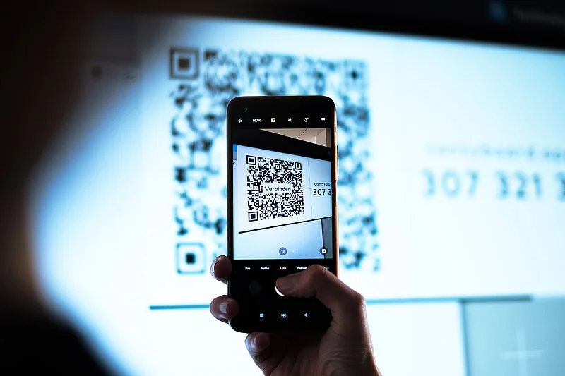 Eine Person scannt einen QR-Code mit einem Smartphone, der auf einem Bildschirm angezeigt wird.