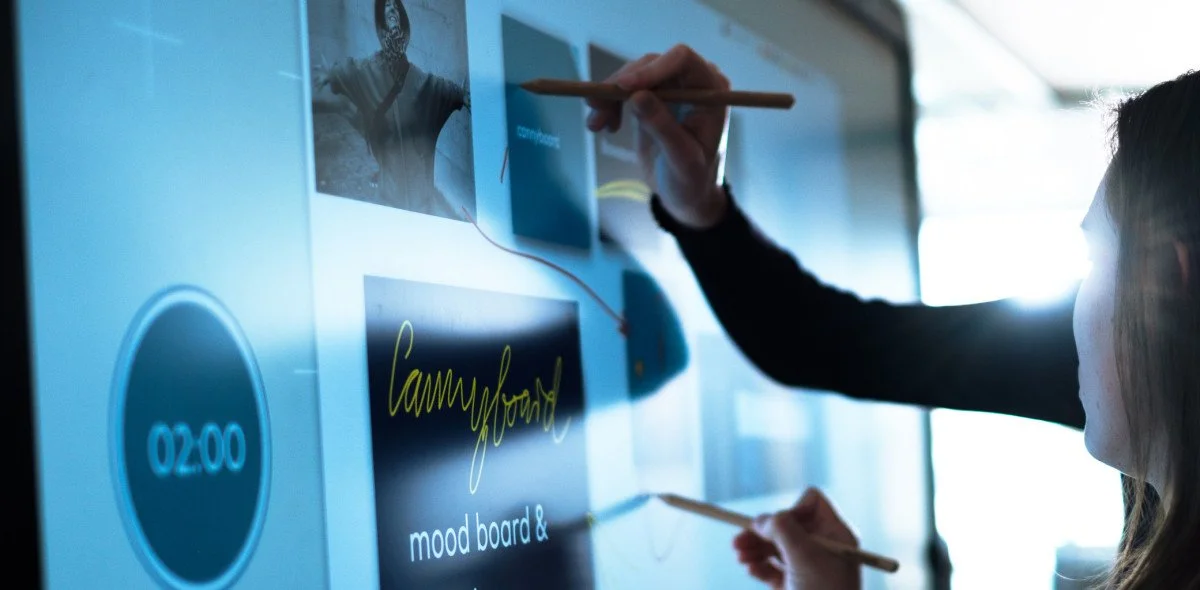 Zwei Personen interagieren mit einem digitalen Wandbild, das ein Kampffeld, eine Uhr und eine Notiz mit der Aufschrift 'mood board' zeigt, in einem modernen Büro.