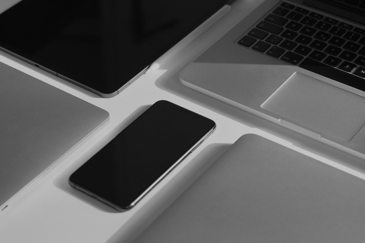 Schwarzes Smartphone liegt auf einem weißen Tisch neben einem Laptop, einem Tablet und einem weiteren elektronischen Gerät.