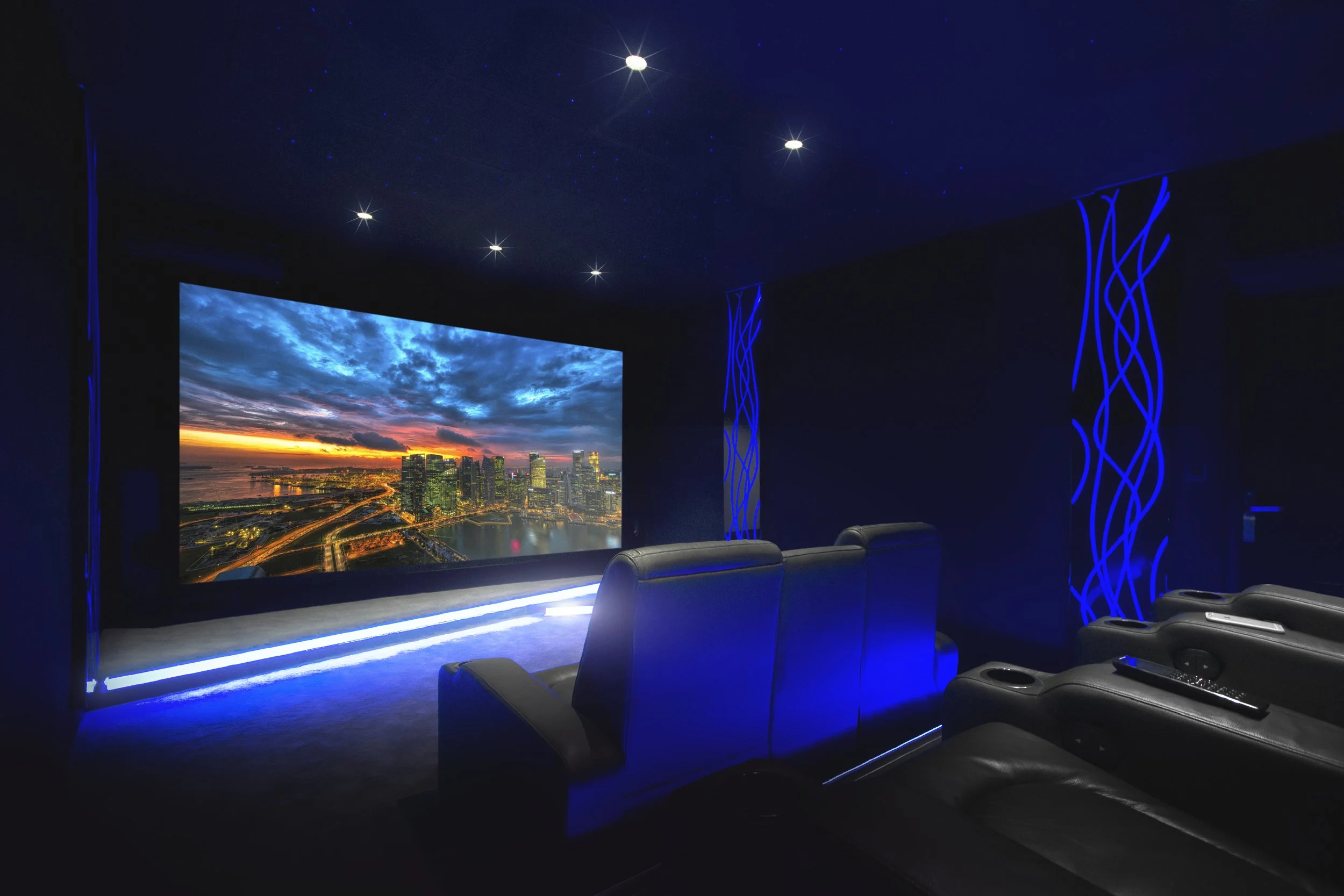 Ein Heimkino mit schwarzen Ledersesseln, einer großen Leinwand mit einem Stadtbild bei Sonnenuntergang und blauer Beleuchtung, umlaufende blaue Lichtleisten und Wanddekoration in dunklen Farben.