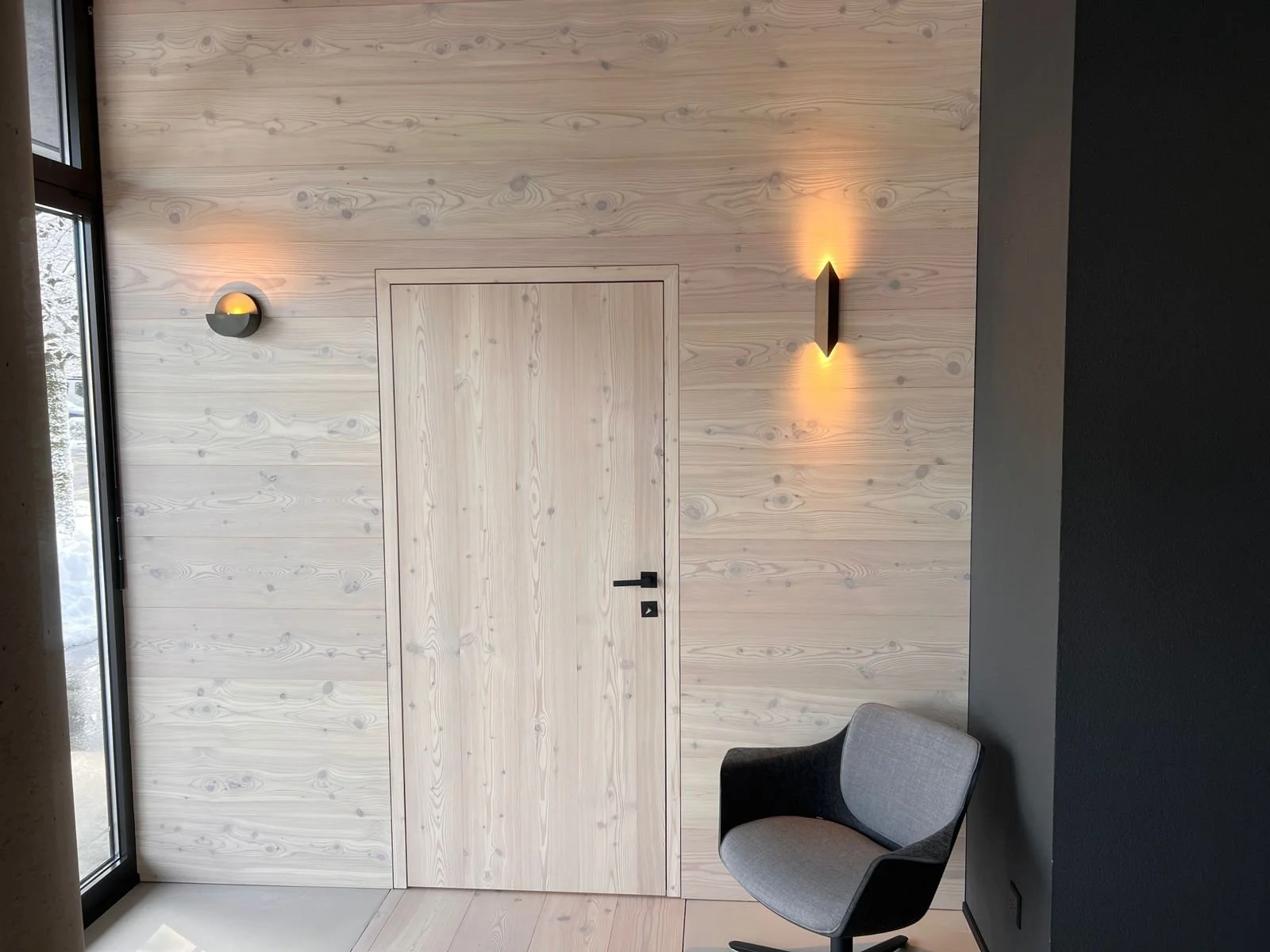 Holzpaneelwand mit schwarzer Wandlampe, graues modernes Sessel, Fenster mit Schnee draußen
