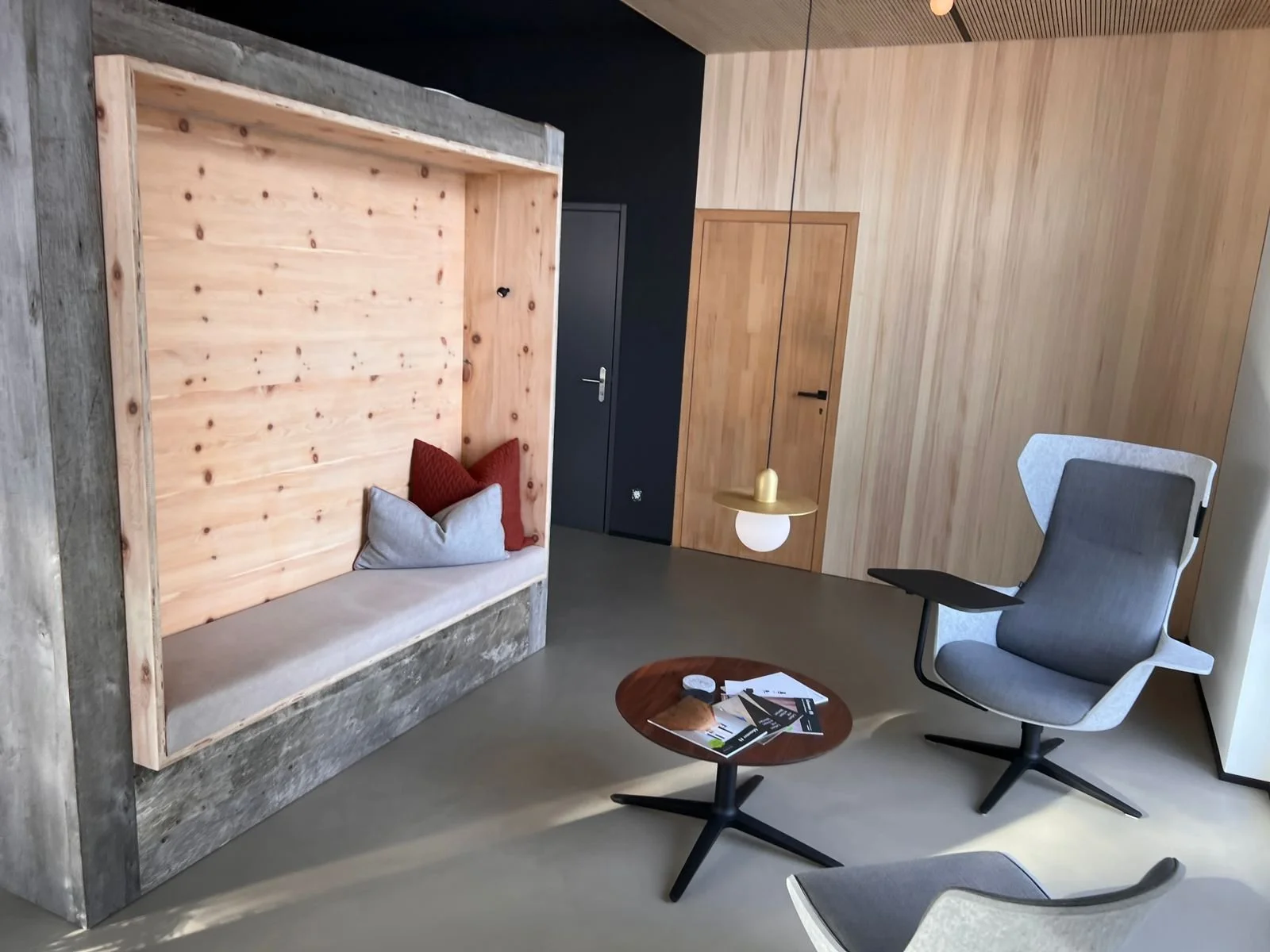 Moderner, minimalistischer Raum mit Holzpaneelen, einem grauen Sessel, einem kleinen runden Tisch mit Zeitschriften und einem besonderen Sitzbereich mit Kissen, einer hängenden Lampe.