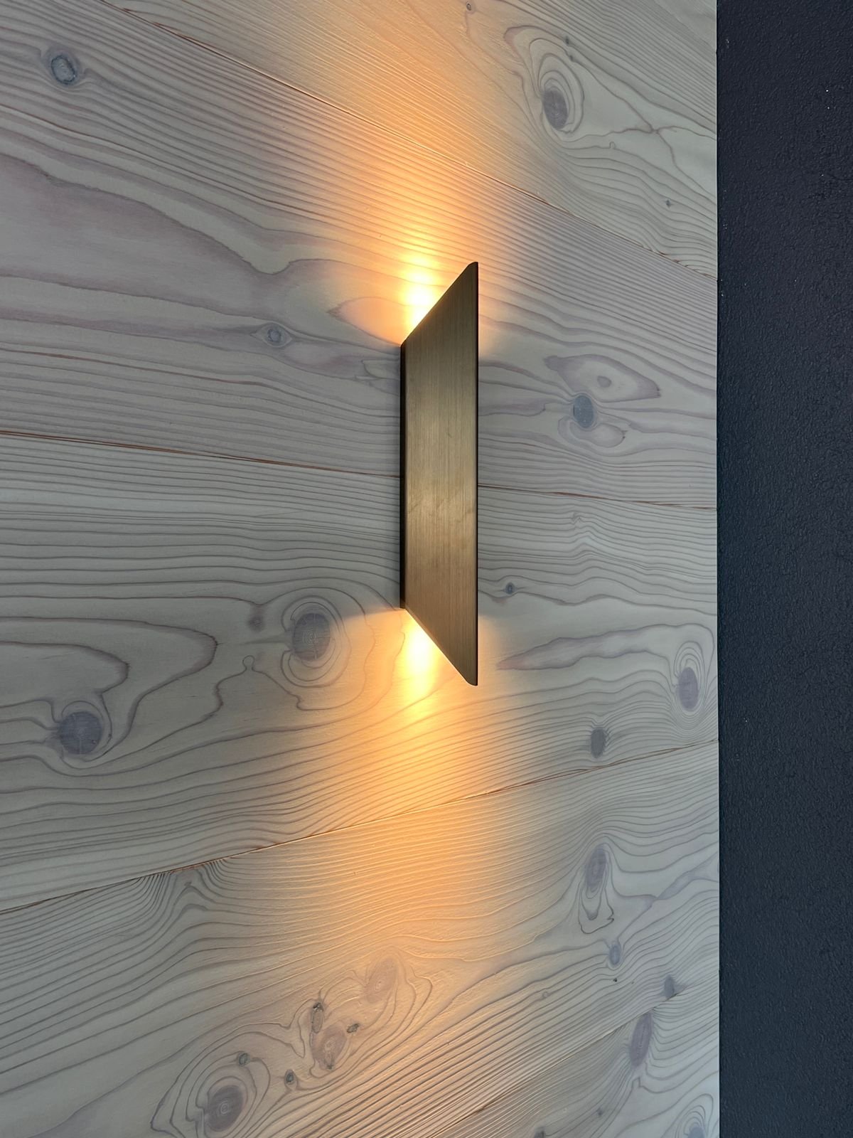 Moderne Wandlampe an einer Holzpaneelwand, die warmes Licht nach oben und unten abstrahlt.