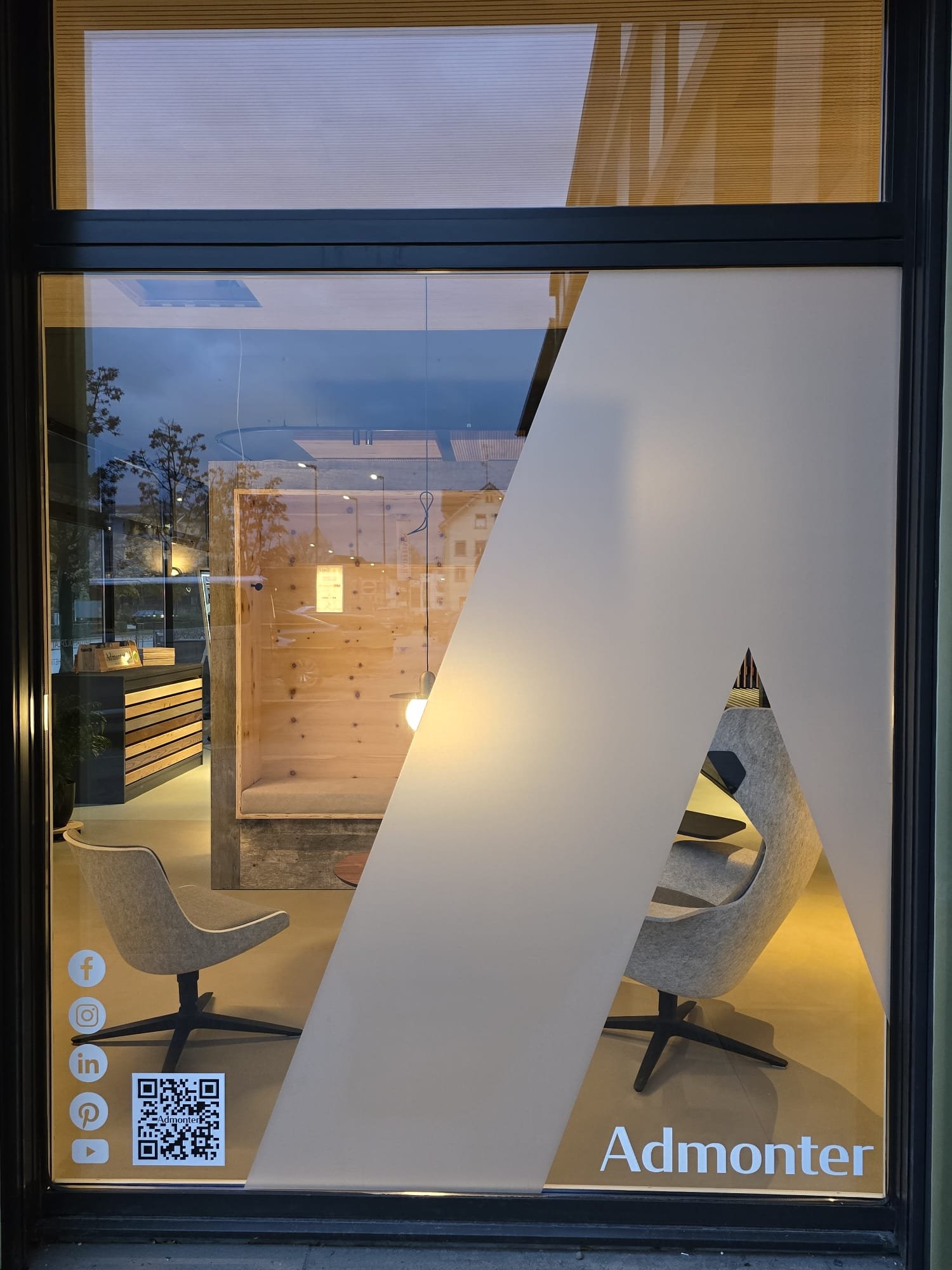 Fensterdeko eines modernen, gemütlichen Bürozimmers mit zwei bequemen Stühlen, einer Stehlampe, Holzelementen und Firmenlogo 'Admonter' im unteren rechten Eck.