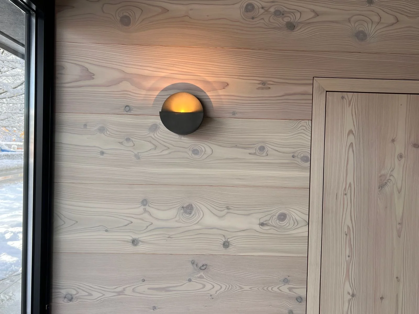 Holzwand mit modernem runden Wandlampen, das Licht ist eingeschaltet, neben einem Fenster, das Schnee im Außenbereich sichtbar macht.