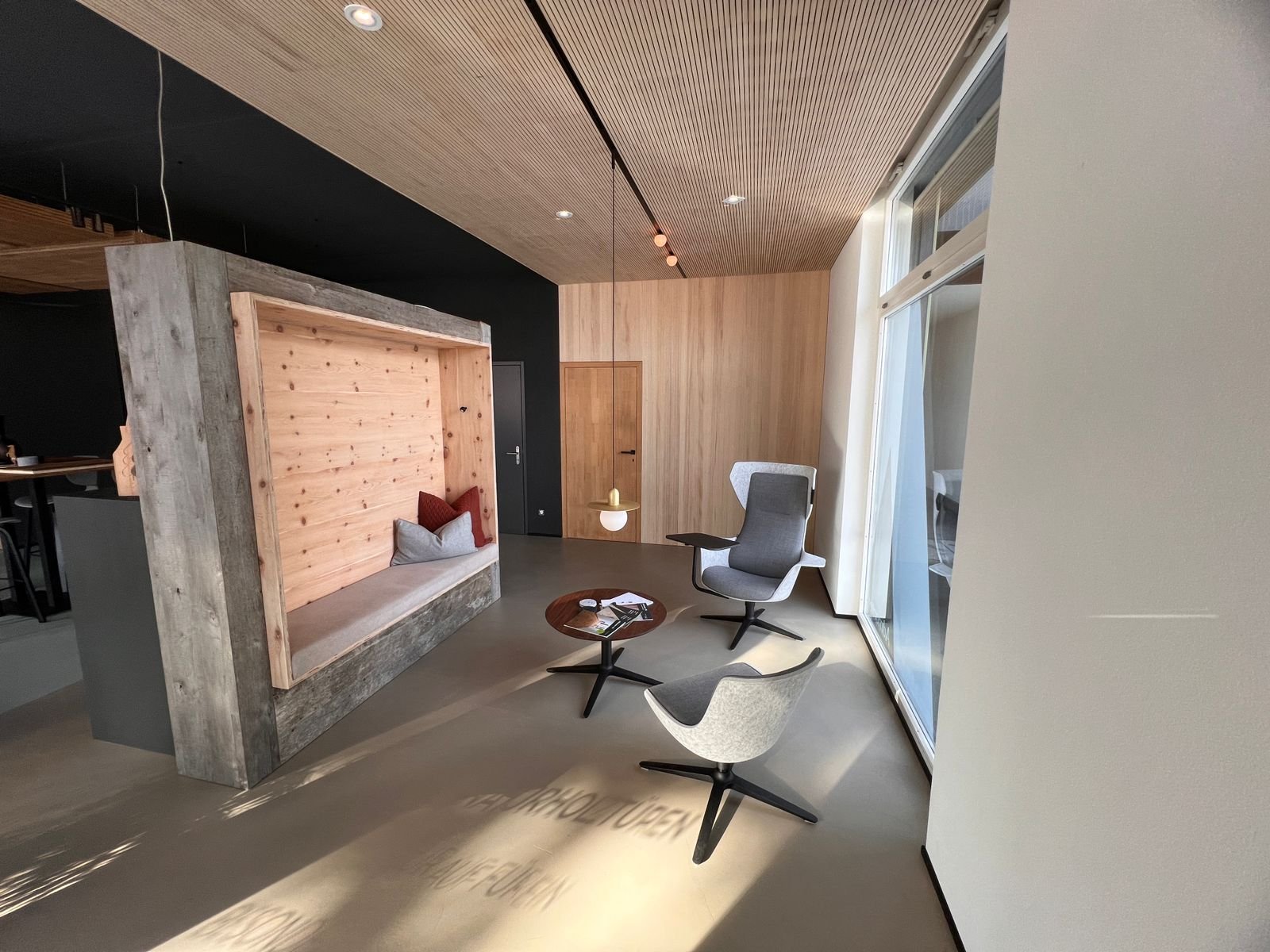 Modernes Wohnzimmer mit Sitzecke, zwei graue Sessel, kleinem runden Tisch, große Fenster, Holzverkleidung und dunkle Wand