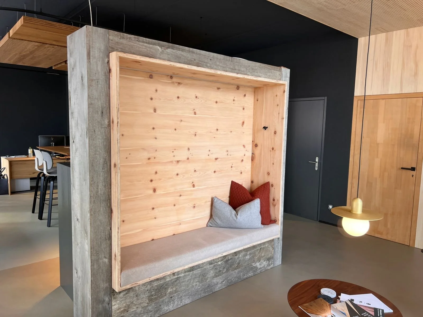 Moderner, offener Sitzbereich mit Kissen, im Hintergrund ein Raum mit Esstischen und Stühlen, moderne Beleuchtung, Holz- und Schwarztöne, Glastisch mit Zeitschriften und Gegenständen.