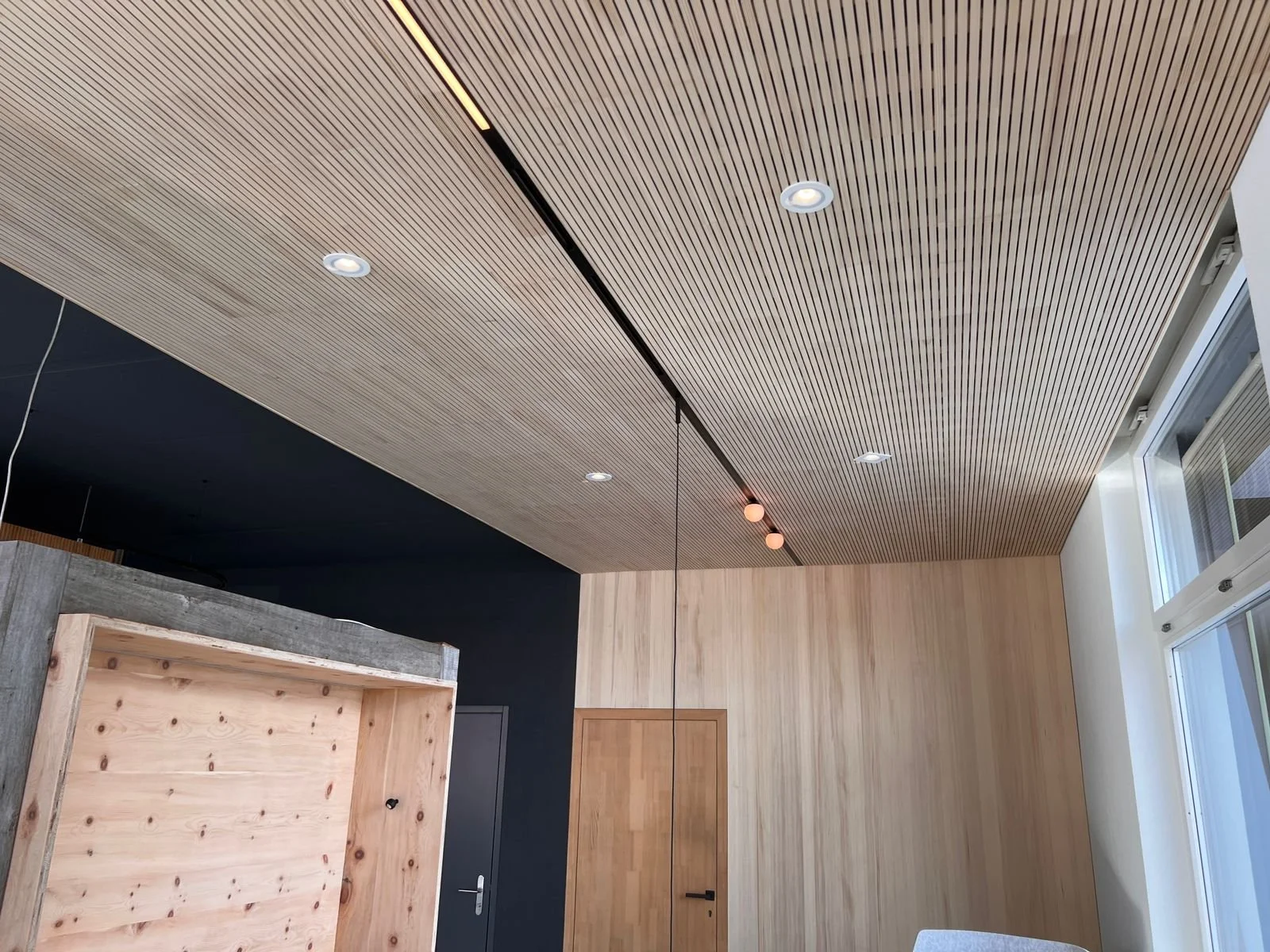 Innenansicht eines Raumes mit Holzboden, Holzdeckenpaneelen, schwarzen Wandflächen, einem Fenster und einer Tür.