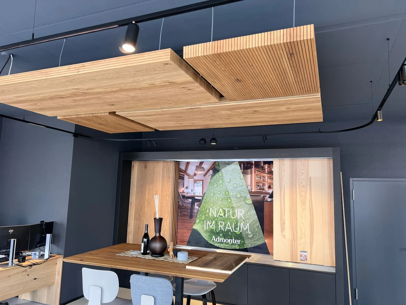 Innenansicht eines modernen Raumes mit dunklen Wänden, Holztischen und -elementen, dekorativen Vasen auf einem Tisch, ein Computer und ein großes Fenster mit einer Werbung für einen Naturraum.