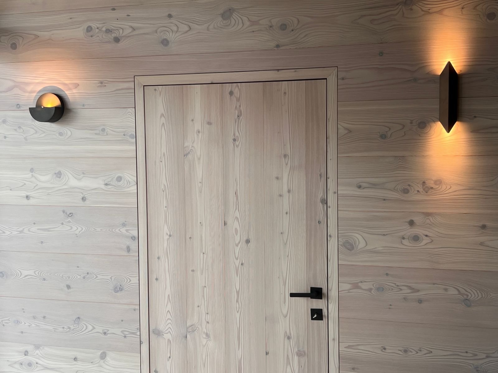 Holztür in einer Wand, umgeben von hellen Holzpaneelen, mit schwarzen Türgriffen und modernen Wandleuchten an beiden Seiten, die warmes Licht abstrahlen.