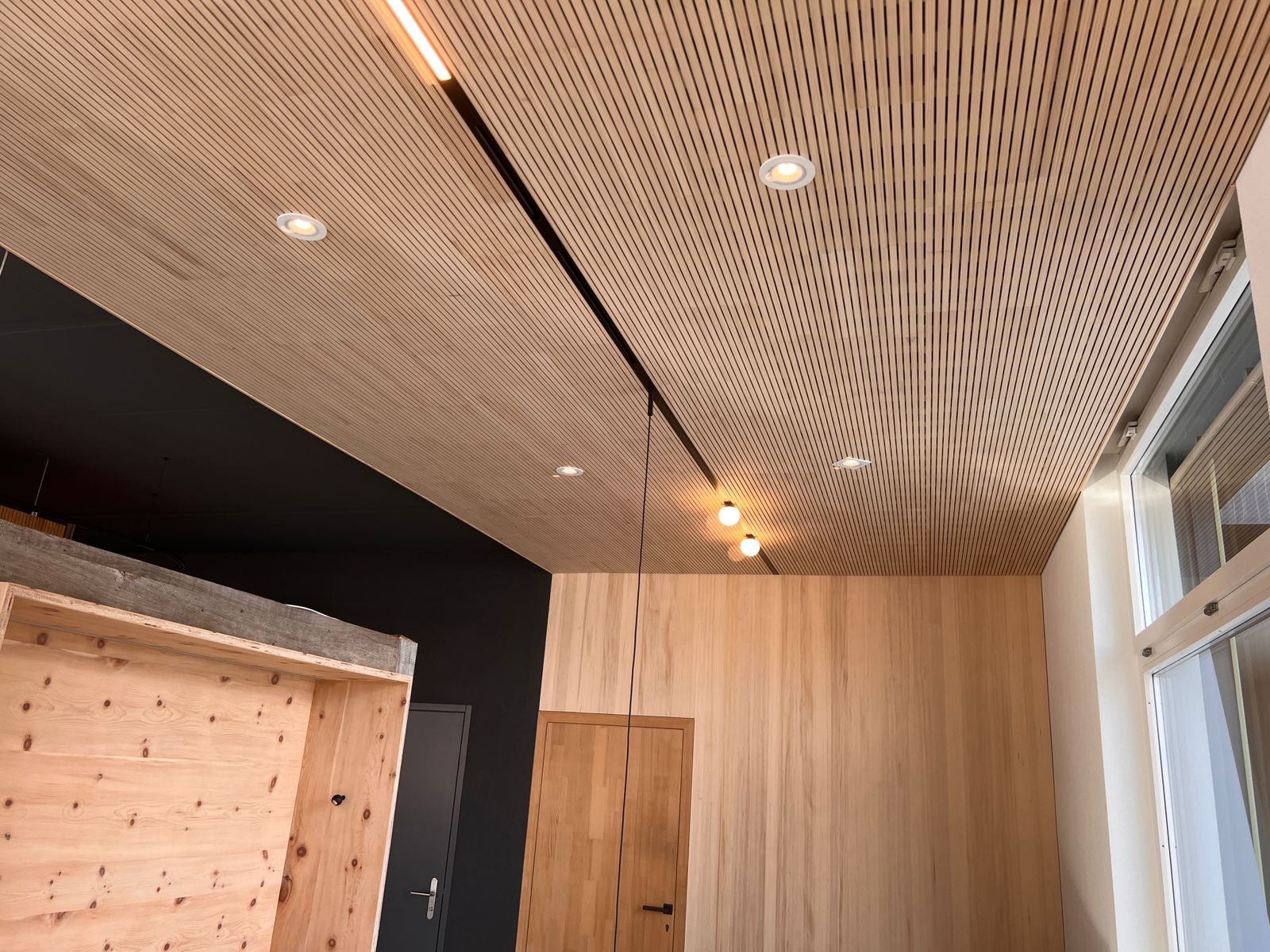 Innenraum mit gemusterter Holzdecke, Holzpaneelen an den Wänden, Fenster und eingebauten Deckenleuchten.