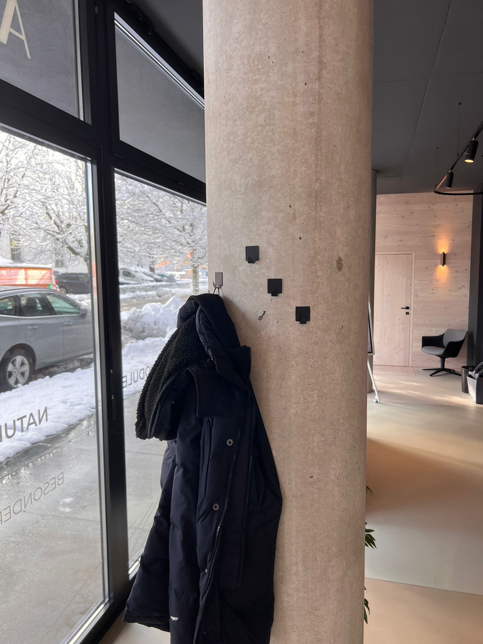 Ein Mantel hängt an einer Wand vor einem Fenster, im Hintergrund sind Schneemenge draußen sichtbar.