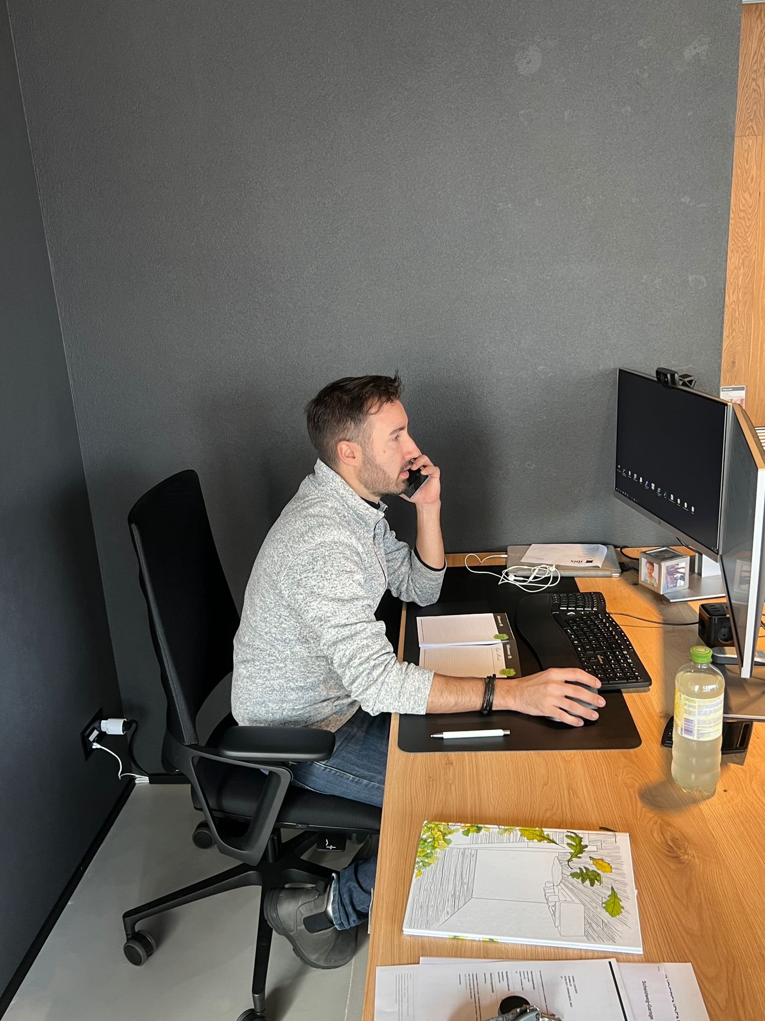 Ein Mann sitzt an einem Büroarbeitsplatz, spricht am Telefon, arbeitet am Computer und hat Notizbücher vor sich.