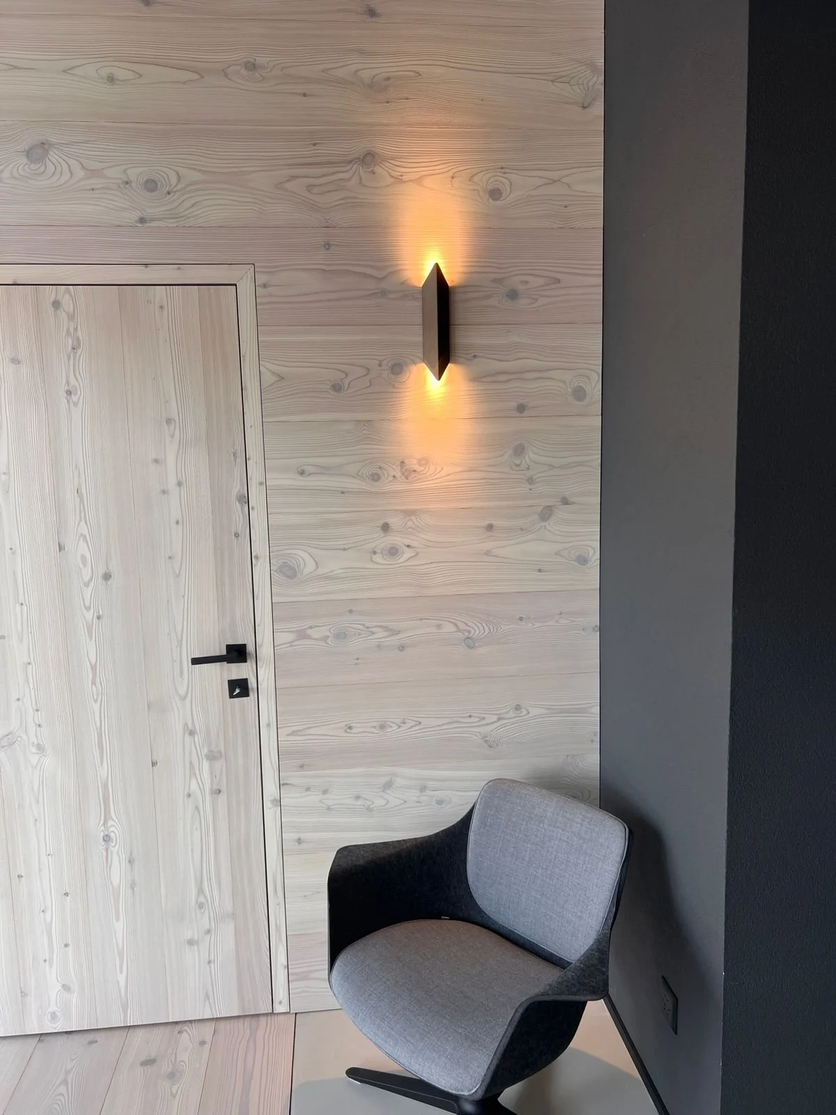 Ein moderner Raum mit einer Wandlampe, einem grauen und schwarzen Sessel, einer Holztür und einer Holz-Wandverkleidung, die einen minimalistischen Stil zeigt.