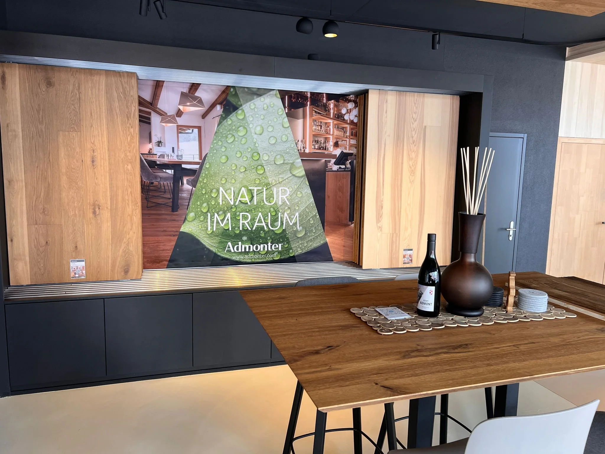 Innenraum mit einem Holzlatztisch, Dekorationsvase und Weinflasche, Blick auf eine große Tür mit einem Poster. Das Poster zeigt einen Raum mit Holzdekor und der Text "Natur im Raum". Darunter steht "Admonter" und eine Website. Der Raum hat dunkle Wän