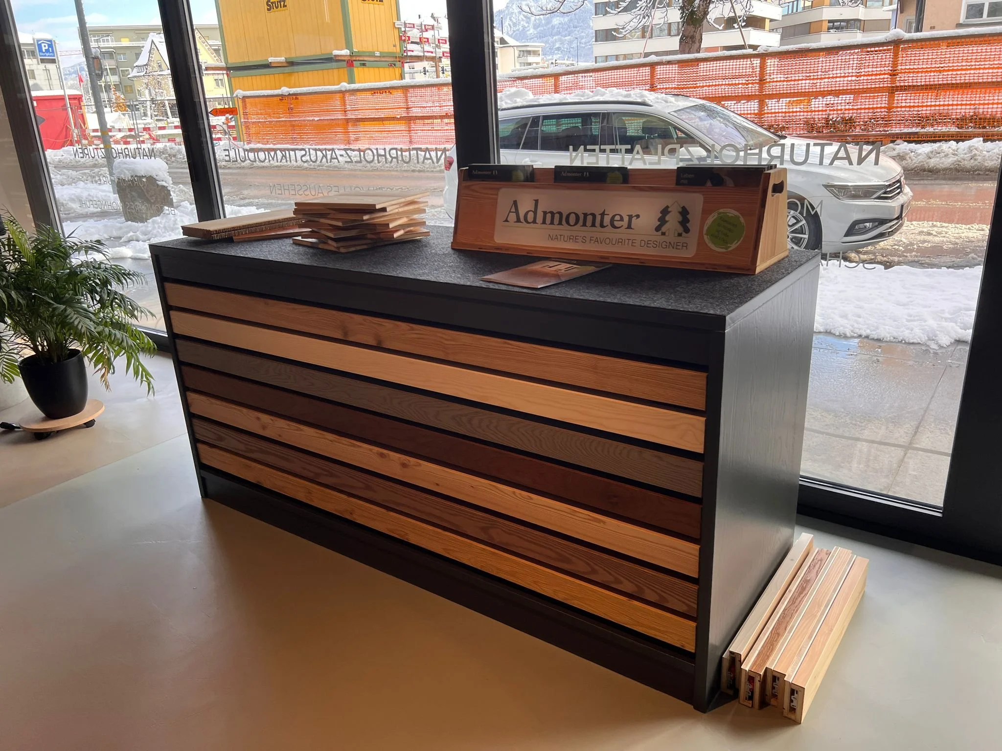 Holzmaterialien und eine Präsentationsbox mit dem Schriftzug "Admonter" auf einer Theke vor einer Glasfront, im Hintergrund Schneeschnee auf dem Boden, ein Auto und Bauarbeiten im Freien.
