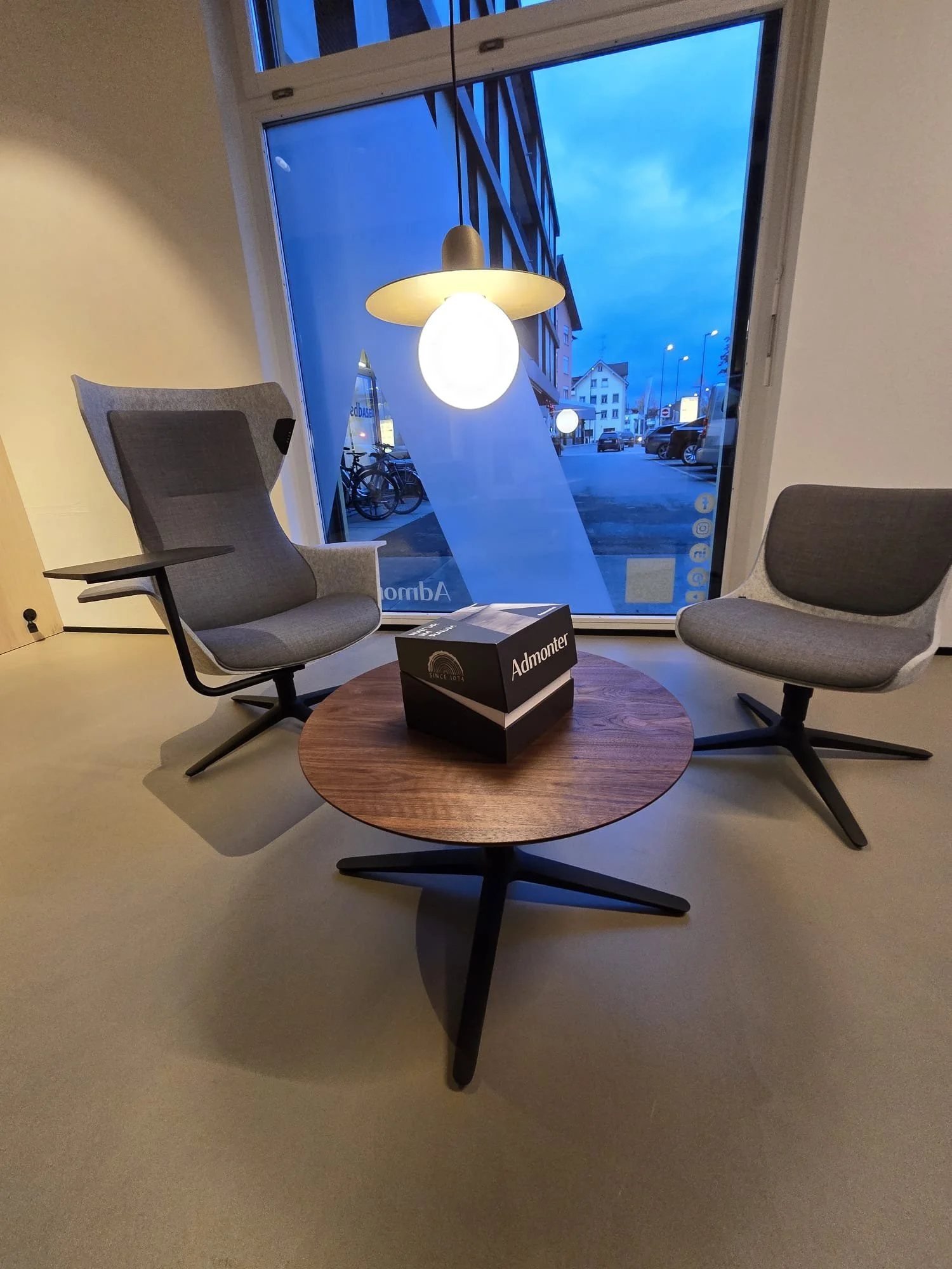 Innenraum mit zwei modernen, grauen, drehbaren Sessel, einem kleinen runden Holztisch und einer pendelnden Deckenlampe. Im Hintergrund eine große Fensterfront mit Blick nach draußen auf eine Straße mit Parkplätzen und Gebäuden bei Dämmerung.