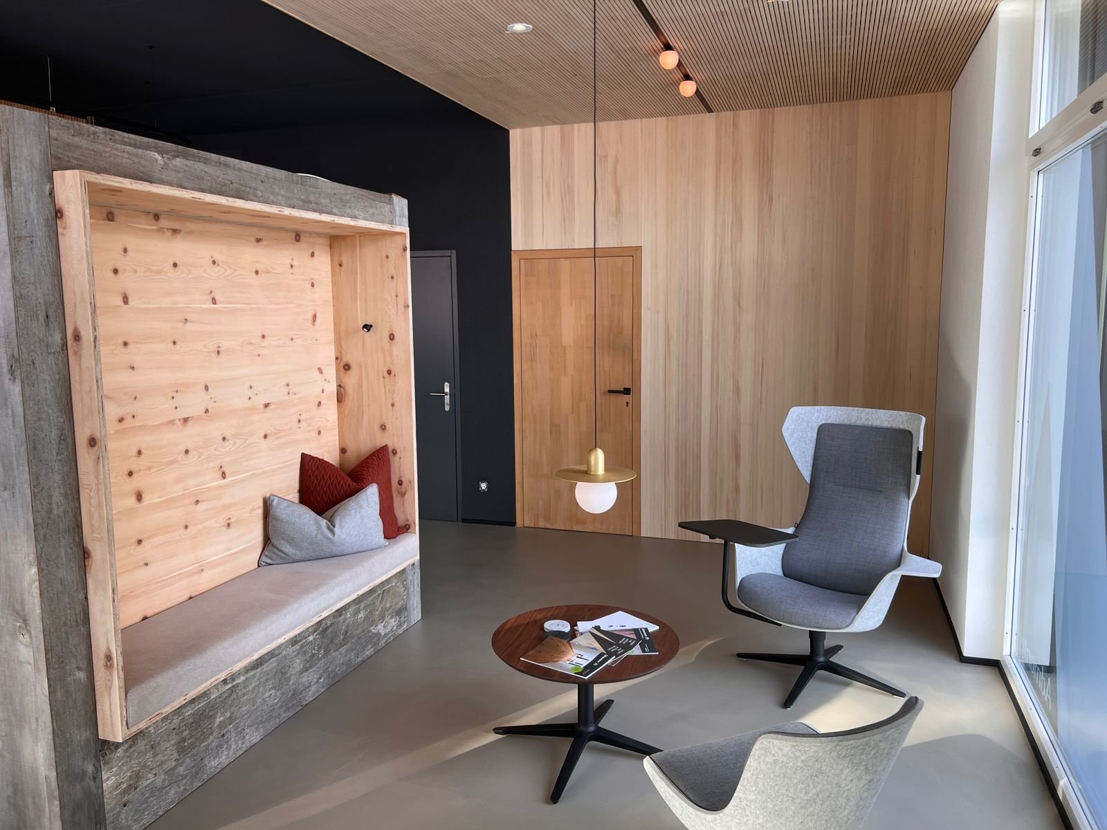 Moderner Sitzbereich mit grauen und schwarzen Sessel, einem runden Holztisch, einem Wandnischen mit Kissen und einem hölzernen Raumteiler, große Fenster an der rechten Seite, Wände aus hellem Holz und eine subtil beleuchtete Decke.