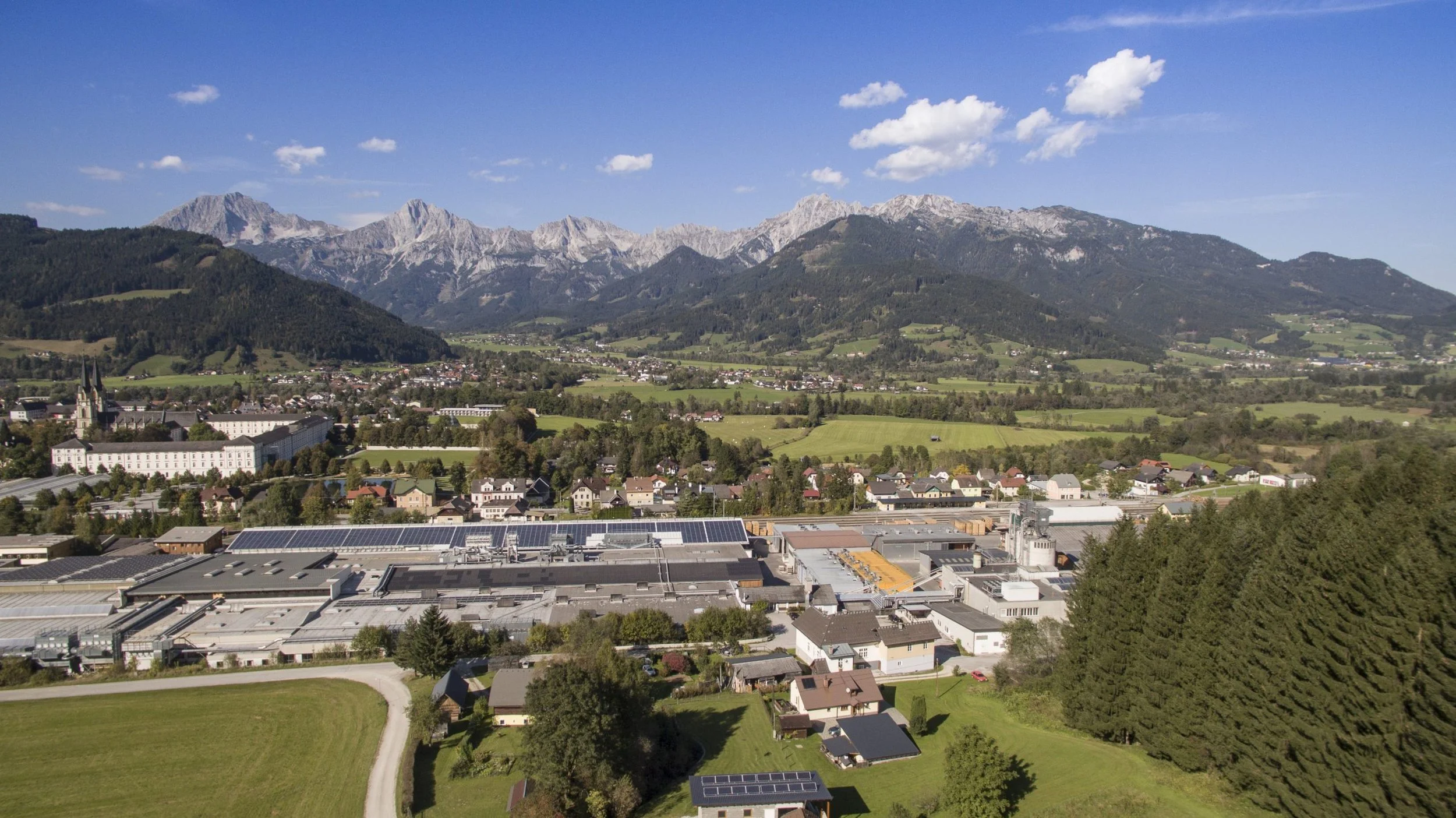 Landschaft mit Berge, Wiesen, Häuser und Fabrik im Vordergrund, blauer Himmel mit Wolken.