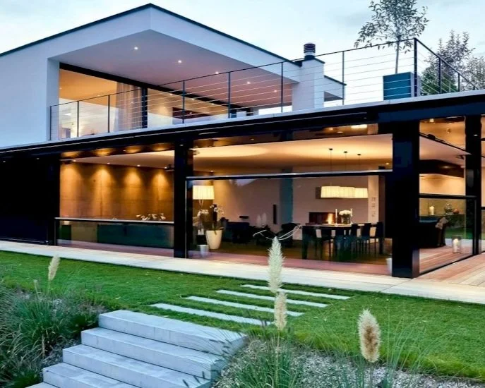 Modernes Haus mit gläserner Veranda, beleuchteter Innenraum, grünem Garten und Steinstufen im Vordergrund.