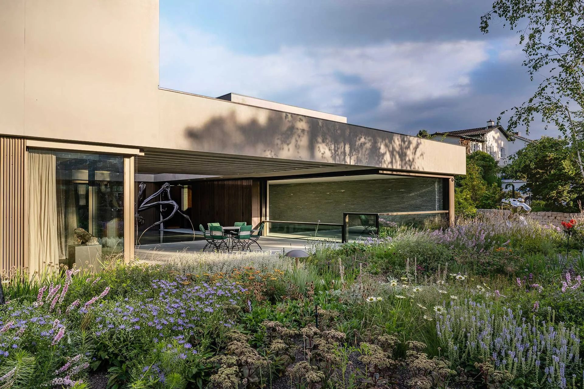 Modernes Haus mit Garten und blühenden Blumen im Vordergrund, sonniger Nachmittag, bewölkter Himmel, Bäume im Hintergrund.