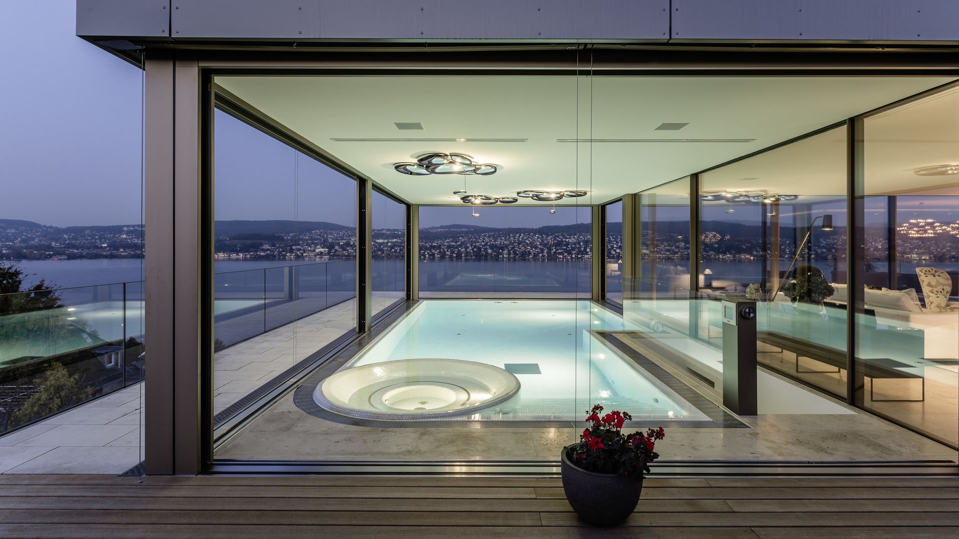 Innenraum eines modernen Hauses mit einem beheizten Pool und Whirlpool, große Glasfenster mit Blick auf eine Stadt bei Abenddämmerung, elegante Beleuchtung und Dekorationen.