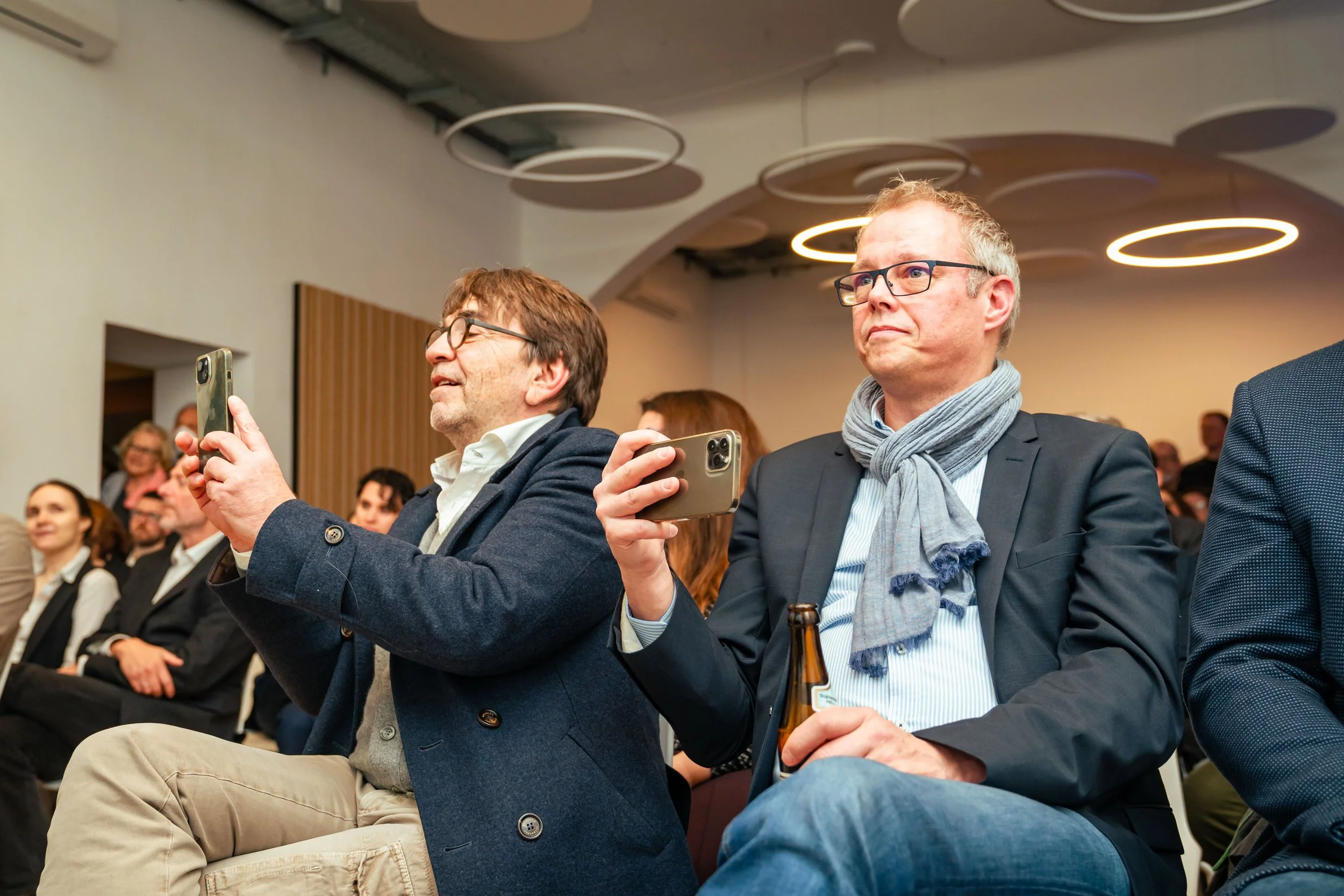 Zwei Männer sitzen in einem Publikum und filmen bei einer Veranstaltung mit ihren Handys, während im Hintergrund weitere Zuschauer sitzen.