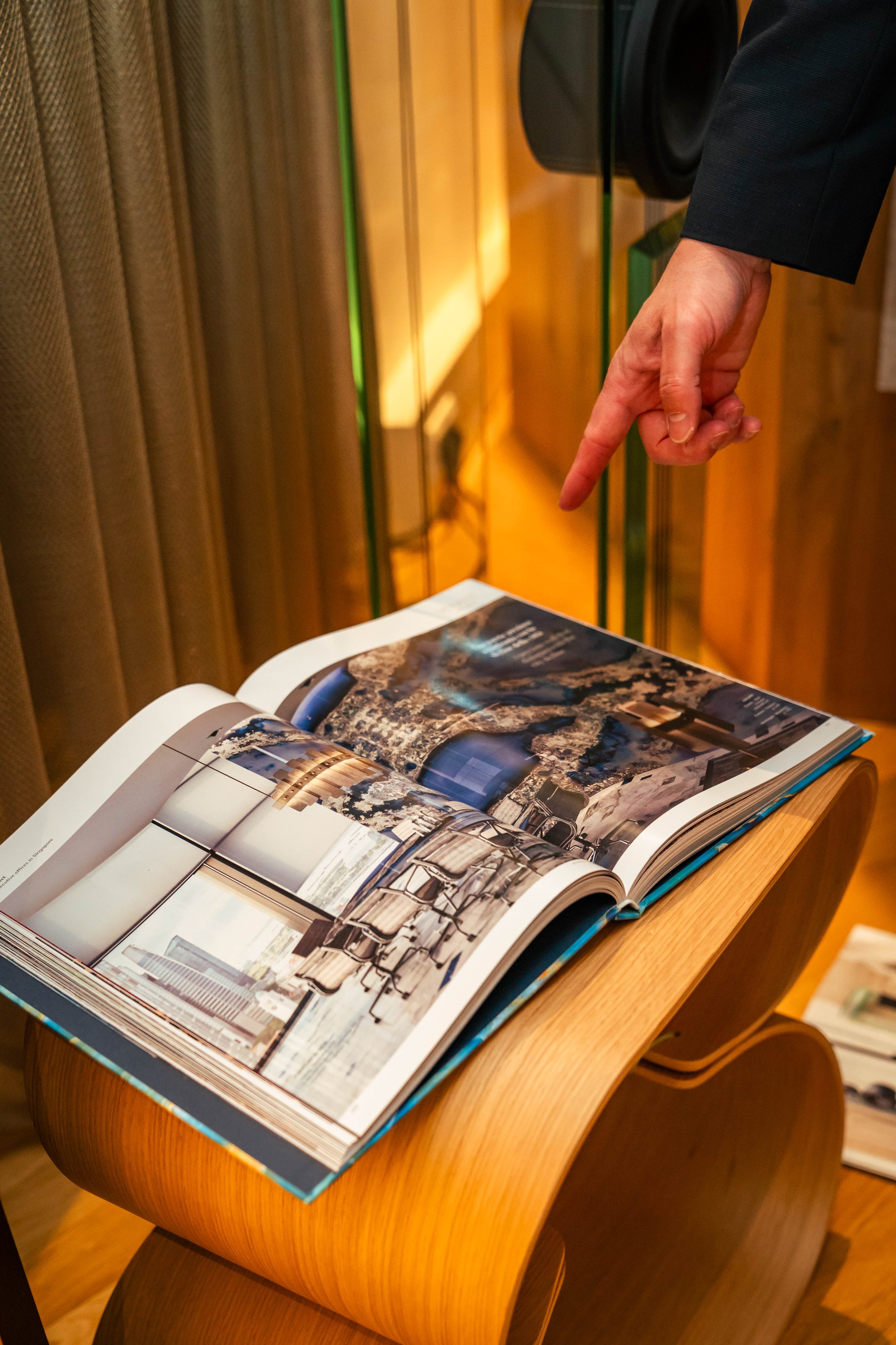 Eine Hand zeigt auf ein großes geöffnetes Fotobuch, das auf einem Holzstativ liegt, mit Gebäuden und Innenarchitekturfotos.