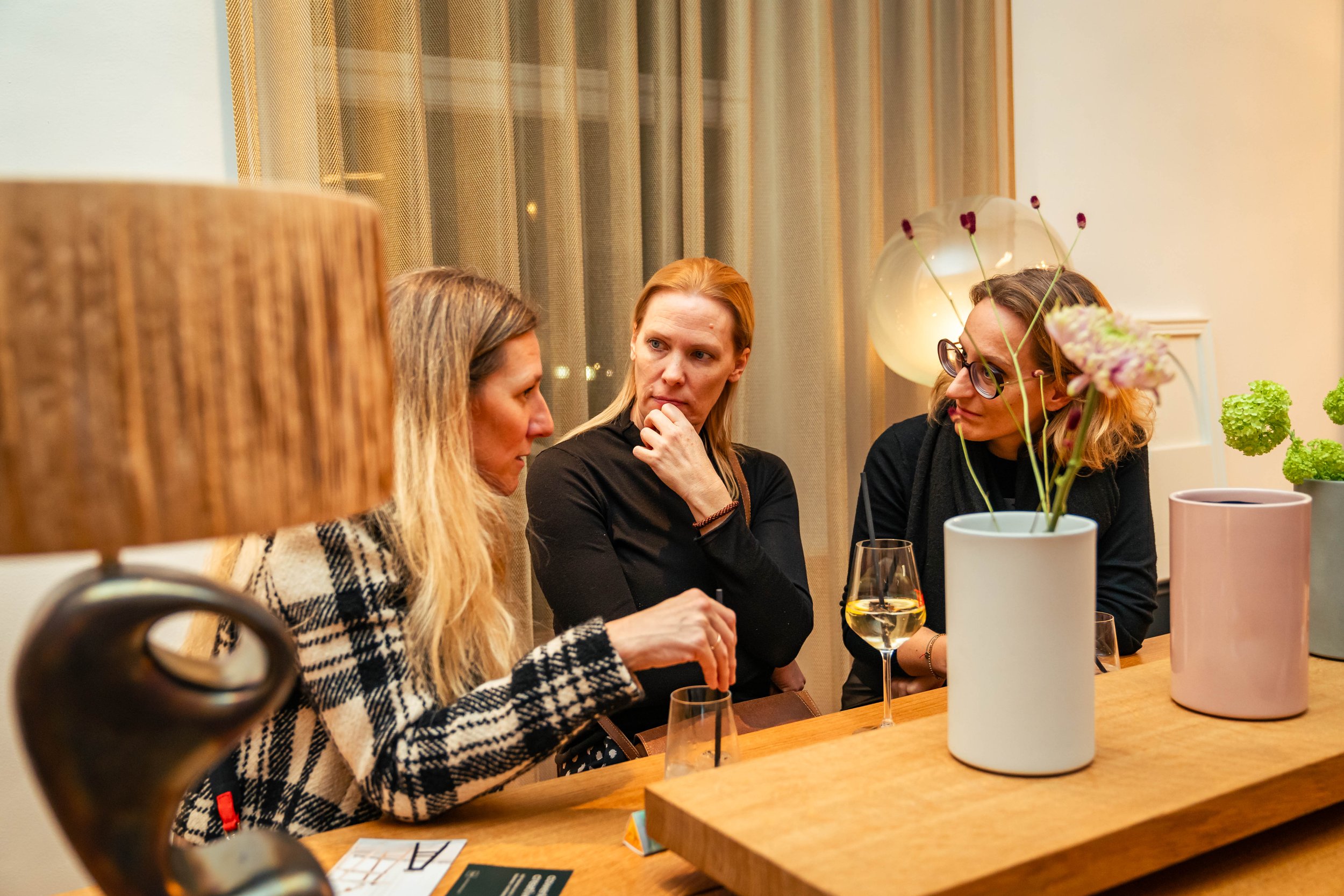 Drei Frauen in einem Gespräch an einem Tisch in einem gemütlichen Raum mit Pflanzen und Lampen.