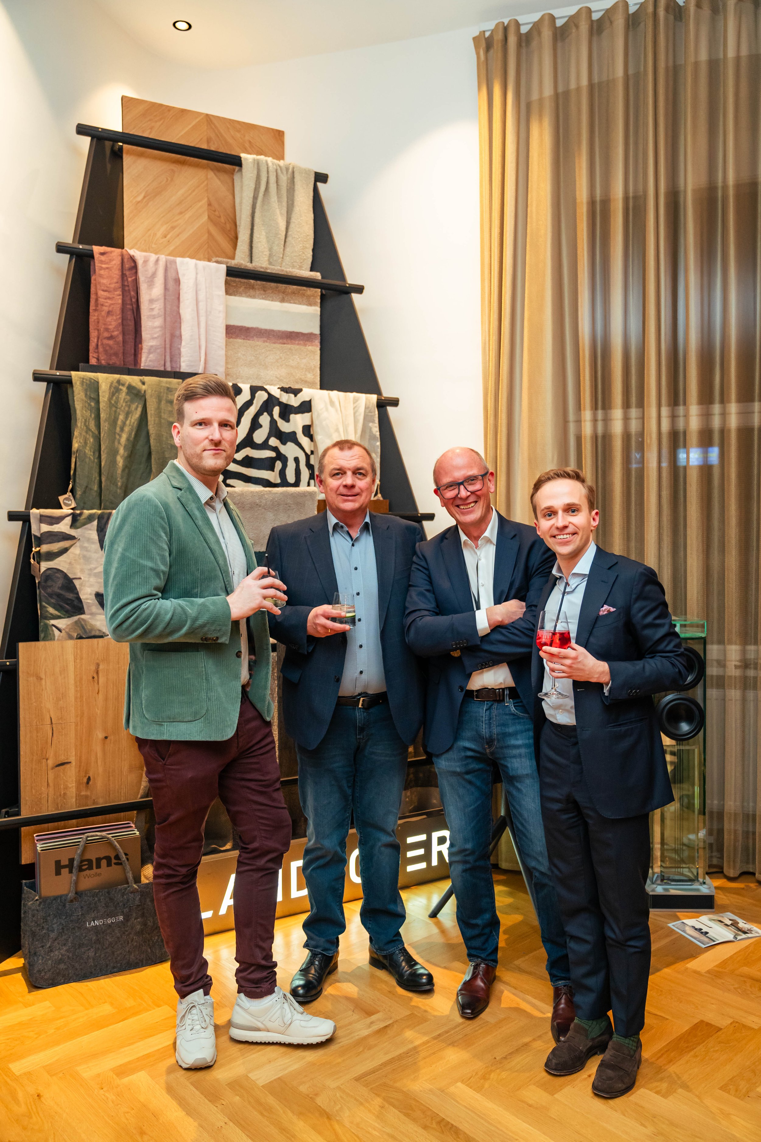 Vier Männer bei einer Veranstaltung im Innenraum, posieren vor einer Wand mit zugeschnittenen Stoffmustern, trinken Wein und lachen. Sie tragen formelle und halbformelle Kleidung. Es gibt große goldene Vorhänge und Holzfußboden im Hintergrund.