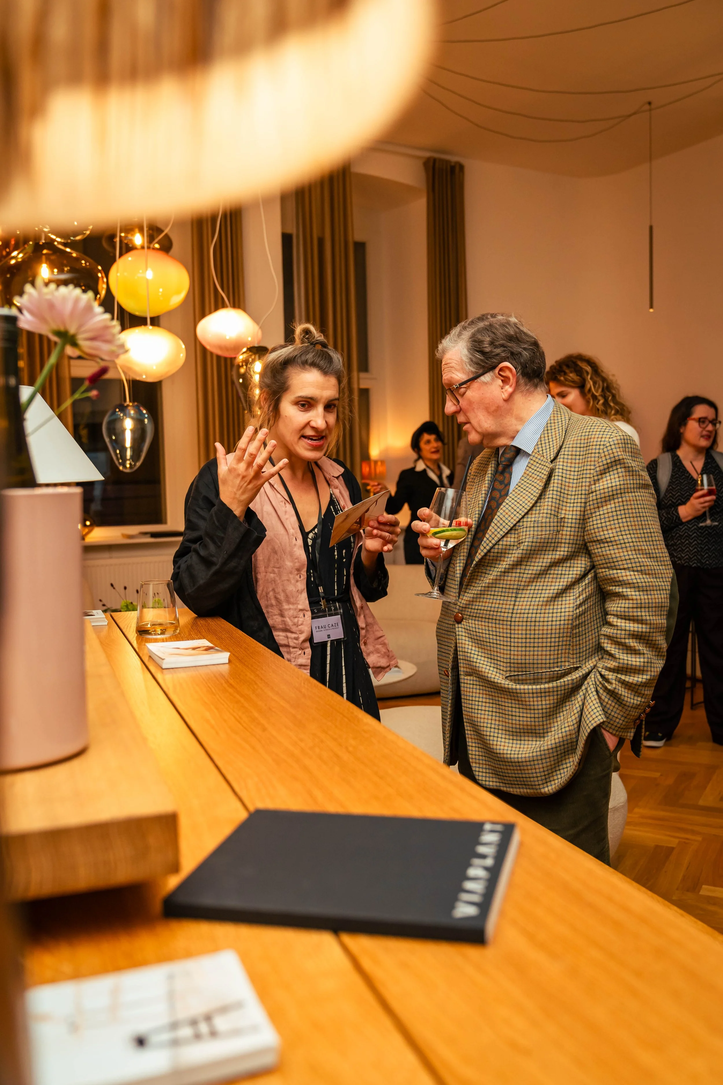 Zwei Menschen unterhalten sich an einer Bar bei einer Veranstaltung, während andere Gäste im Hintergrund sprechen.