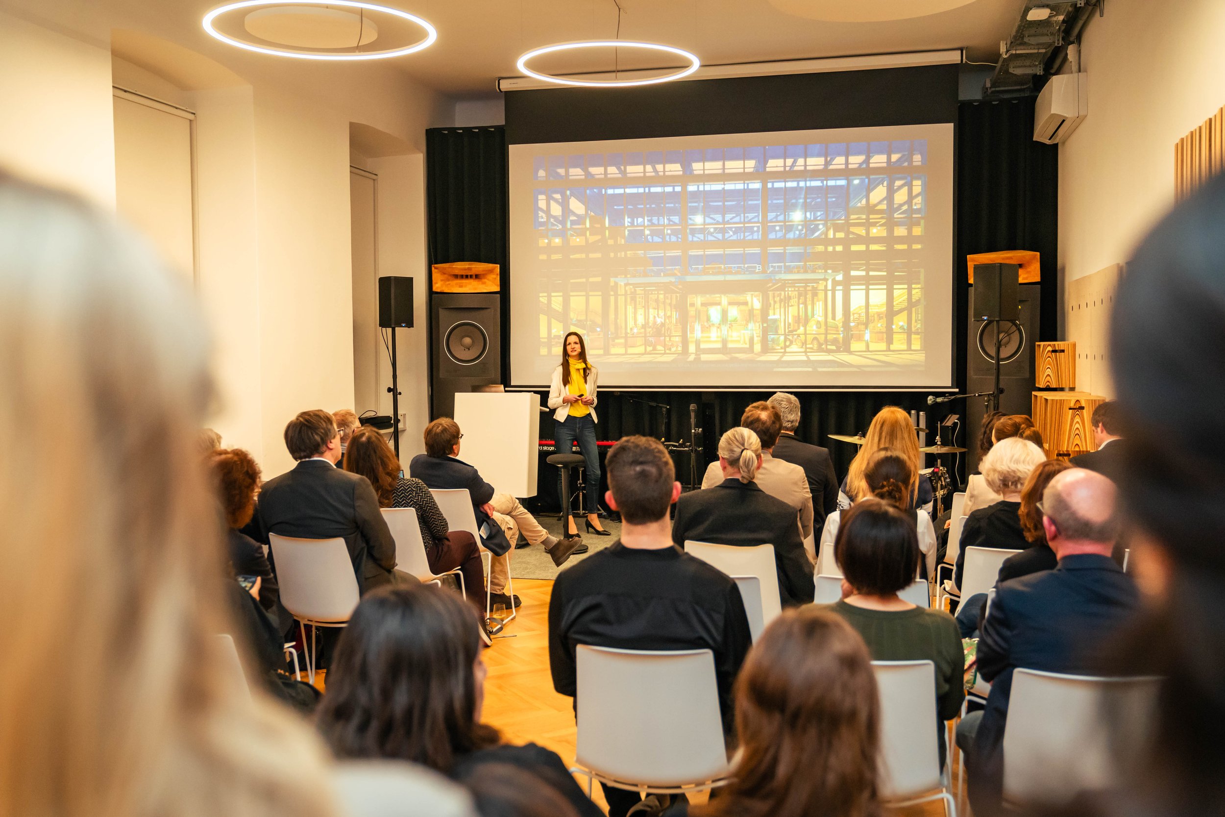Menschen sitzen in einem Auditorium und hören einer Präsentation zu, während die Referentin vor einem Bildschirm steht.