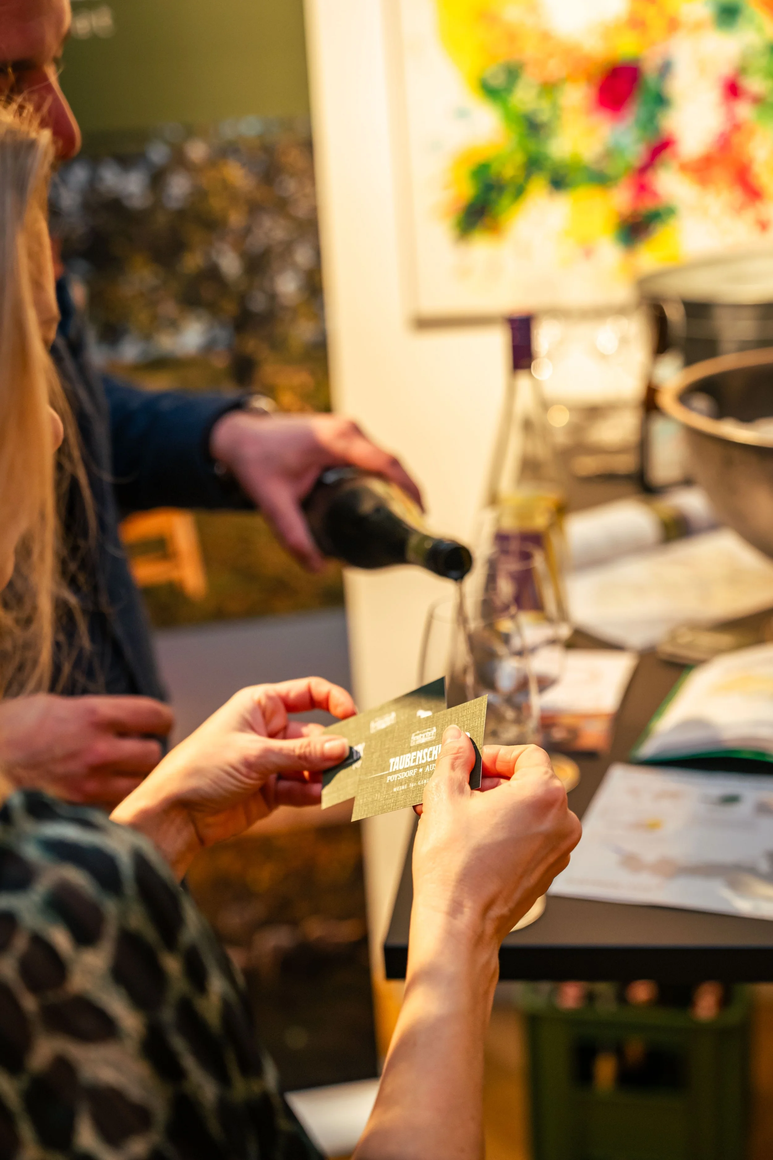 Eine Person hält eine Karte mit Text, während eine andere Person in einem Hintergrund ein Glas mit Getränk einschenkt. Es gibt einen Tisch mit offenen Büchern und Flaschen, im Hintergrund ein farbenfrohes Kunstwerk.