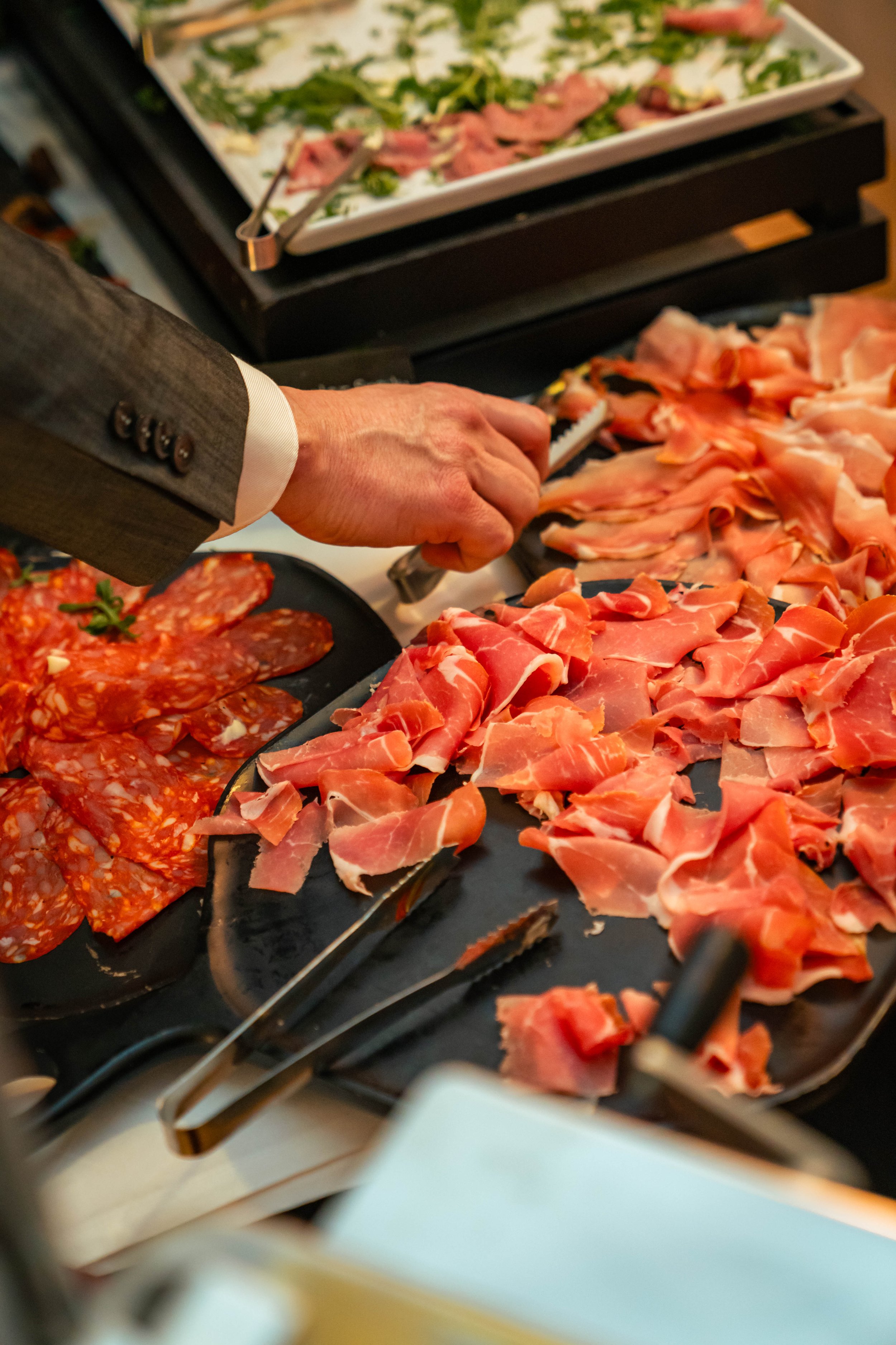 Ein Mann in einem grauen Anzug bedient sich an einer Theke mit verschiedenen Schinken- und Wurstsorten, die auf schwarzen Tellern angerichtet sind, während im Hintergrund ein weiterer Teller mit Salat und rohem Fleisch sichtbar ist.