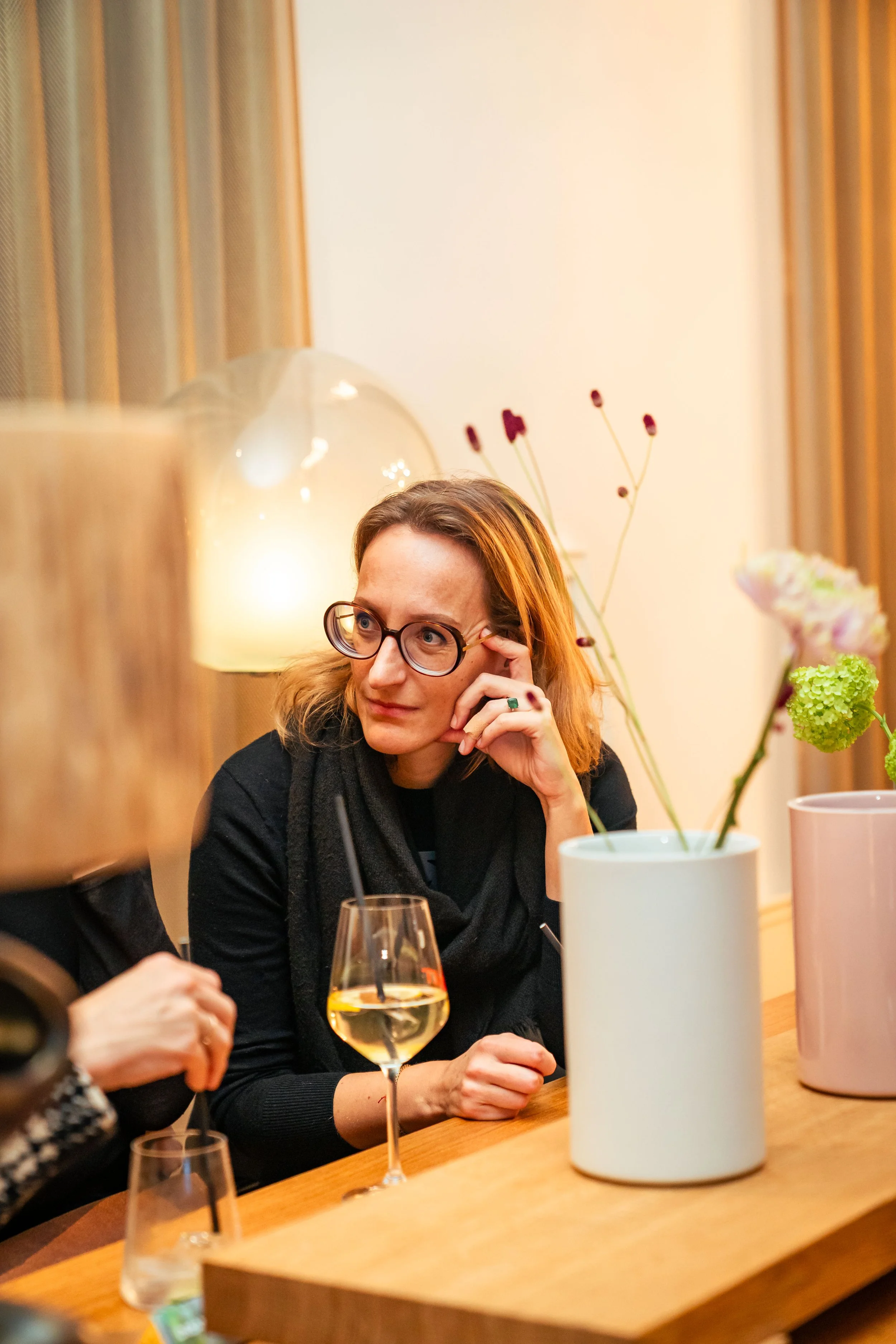 Eine Frau mit Brillen sitzt an einem Tisch mit einer weißen Vase, die Blumen enthält, und einem Glas weißen Weins.