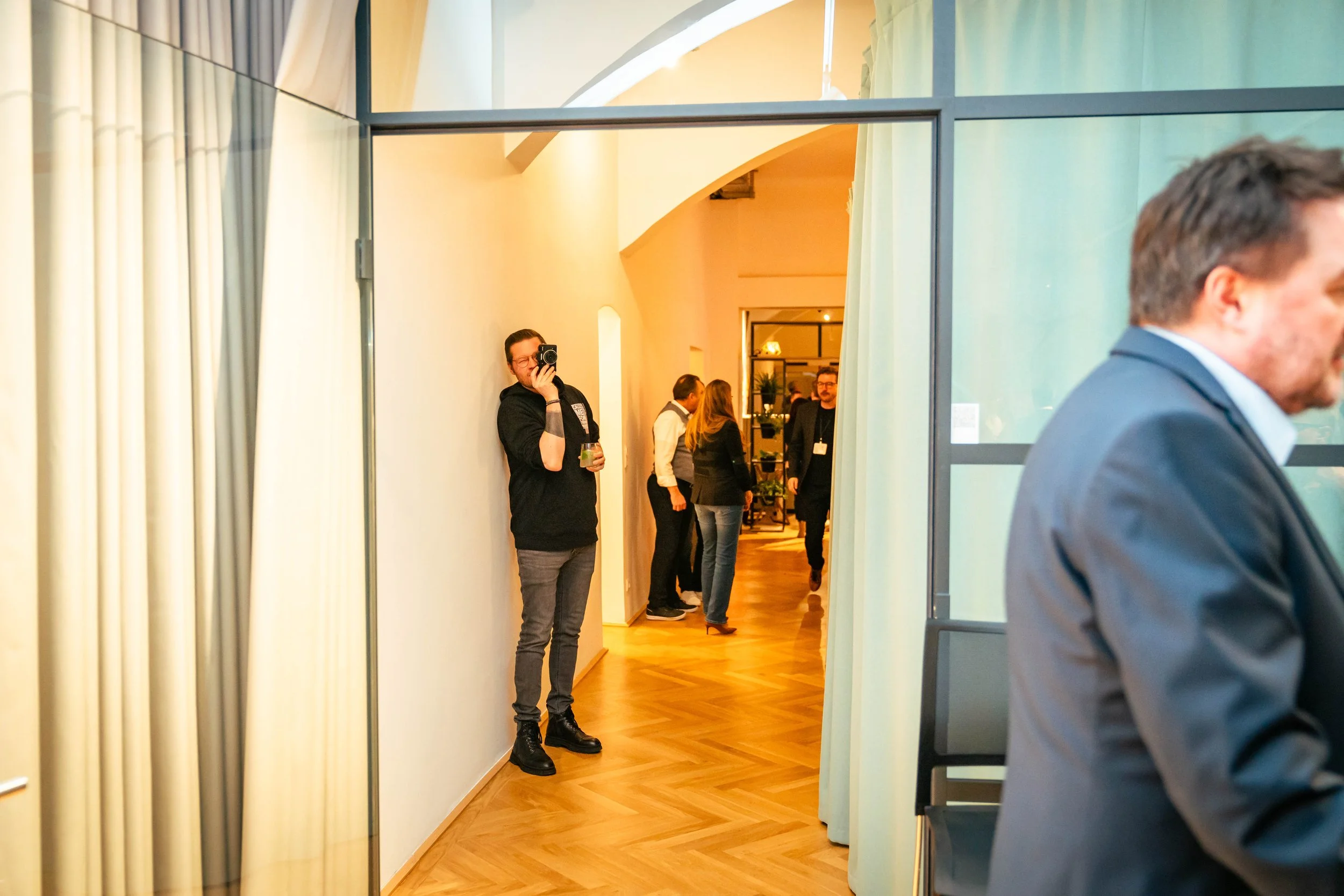 Menschen bei einer Veranstaltung, einige im Hintergrund, eine Person links lehnt an der Wand und fotografiert mit einer Kamera, andere unterhalten sich im Raum, Polsterung und Vorhänge im Raum sichtbar.