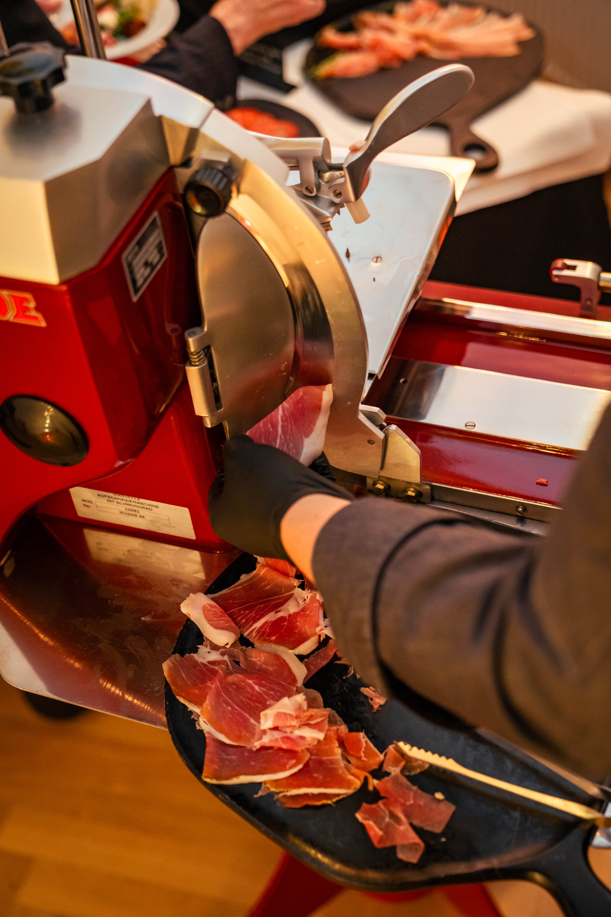 Person schneidet Schinken mit einer Maschine, im Hintergrund sind Platten mit Kaiserschmarrn und andere Speisen auf einem Tisch.