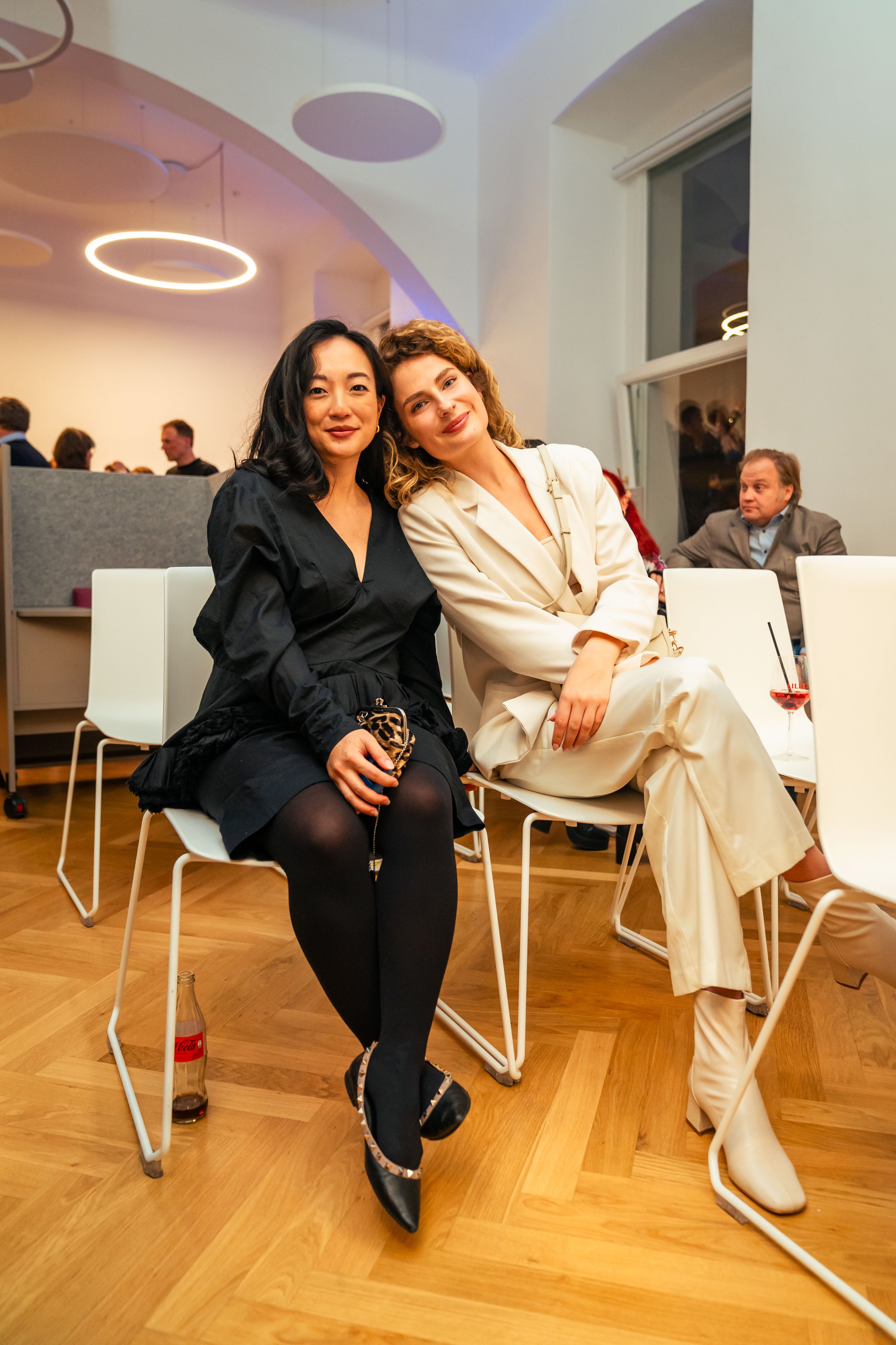 Zwei Frauen sitzen nebeneinander auf Stühlen in einem eleganten Raum, lachen und posieren für die Kamera.