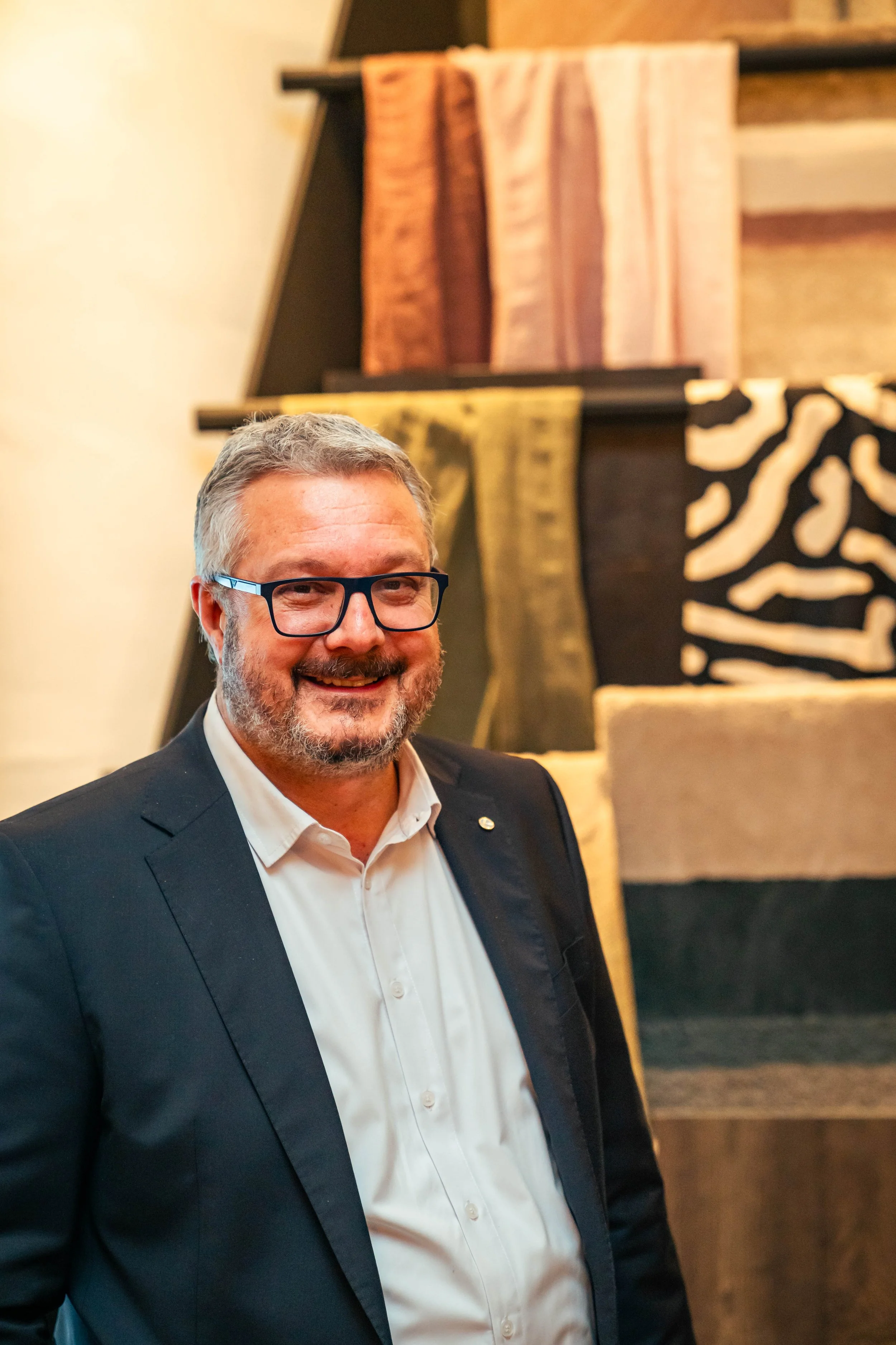 Ein lächelnder Mann mit Brille in formeller Kleidung vor einem Wandbild mit verschiedenen Stoffmustern.