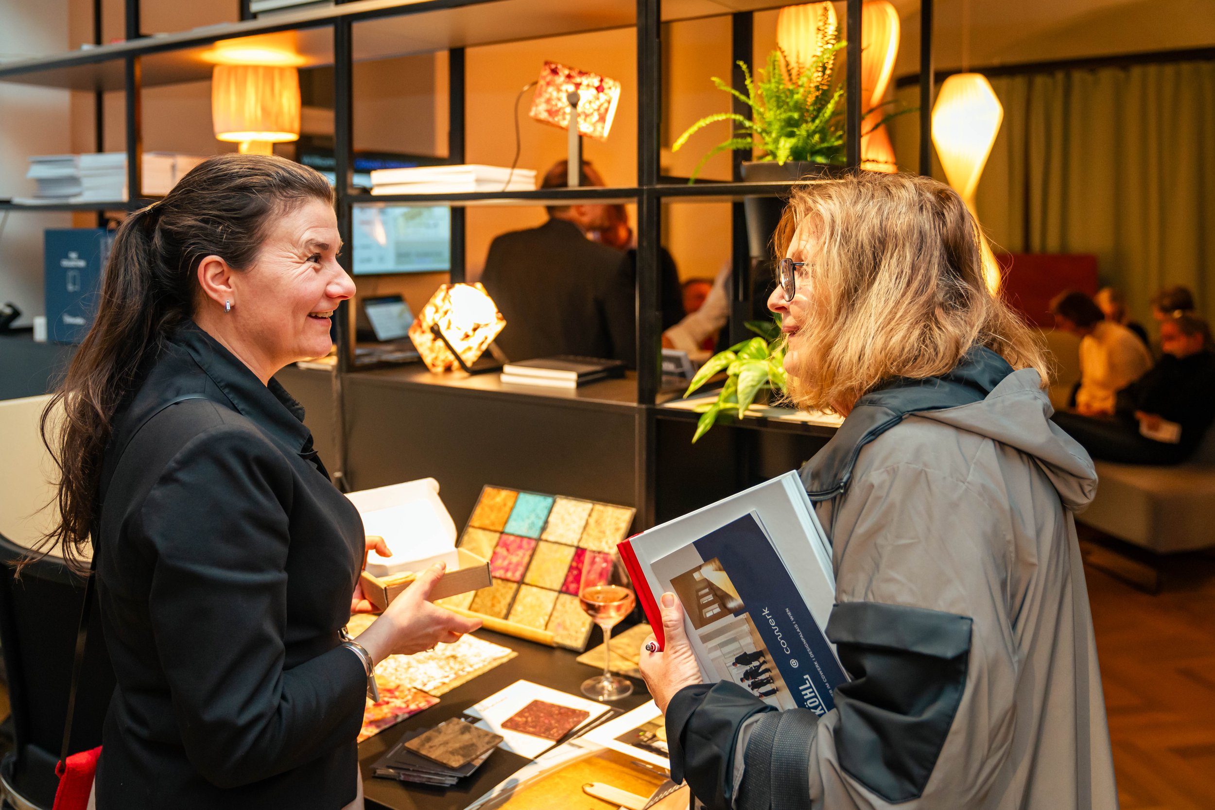 Zwei Frauen sprechen in einem stilvollen Innenraum, eine Verkäuferin zeigt der Kundin Muster und Produkte an einem Verkaufsstand, im Hintergrund sind weitere Menschen und dekorative Lampen.