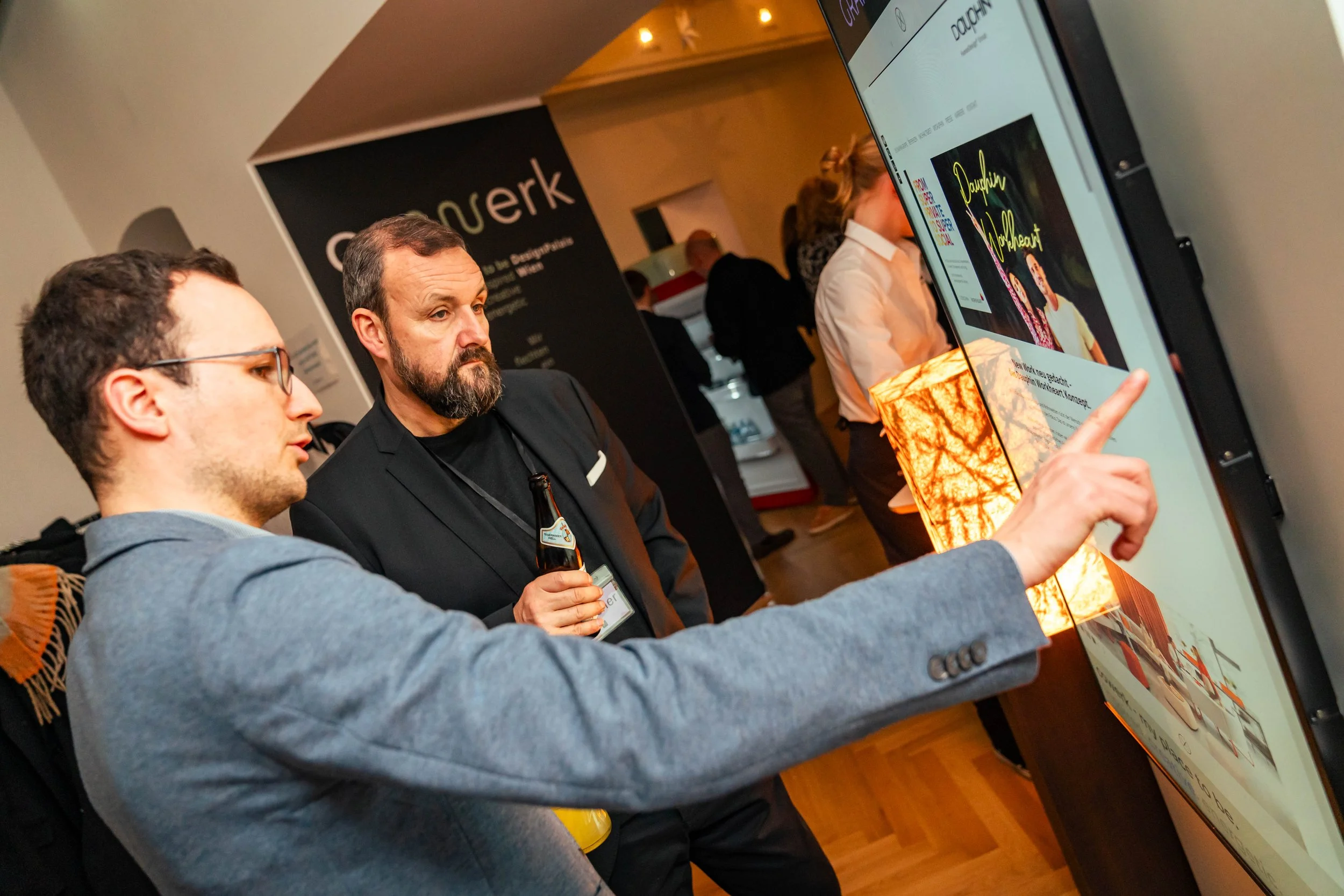 Zwei Männer schauen auf einen digitalen Bildschirm bei einer Veranstaltung. Der eine Mann zeigt auf den Bildschirm, der andere hält ein Getränk in der Hand.
