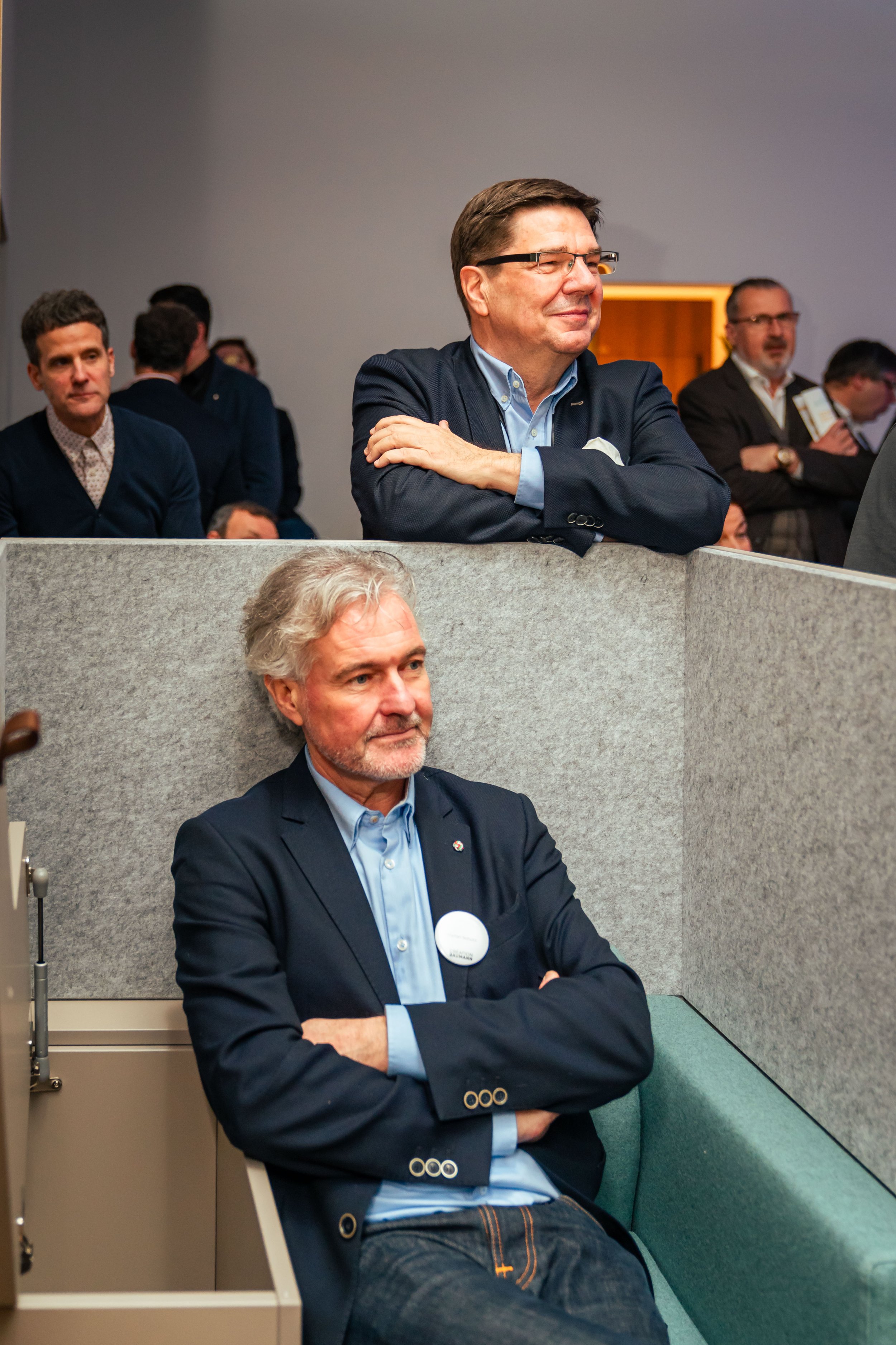 Zwei Männer bei einer Veranstaltung, einer sitzt in einem abgetrennten Bereich, der andere steht daneben, umgeben von weiteren Personen im Hintergrund.