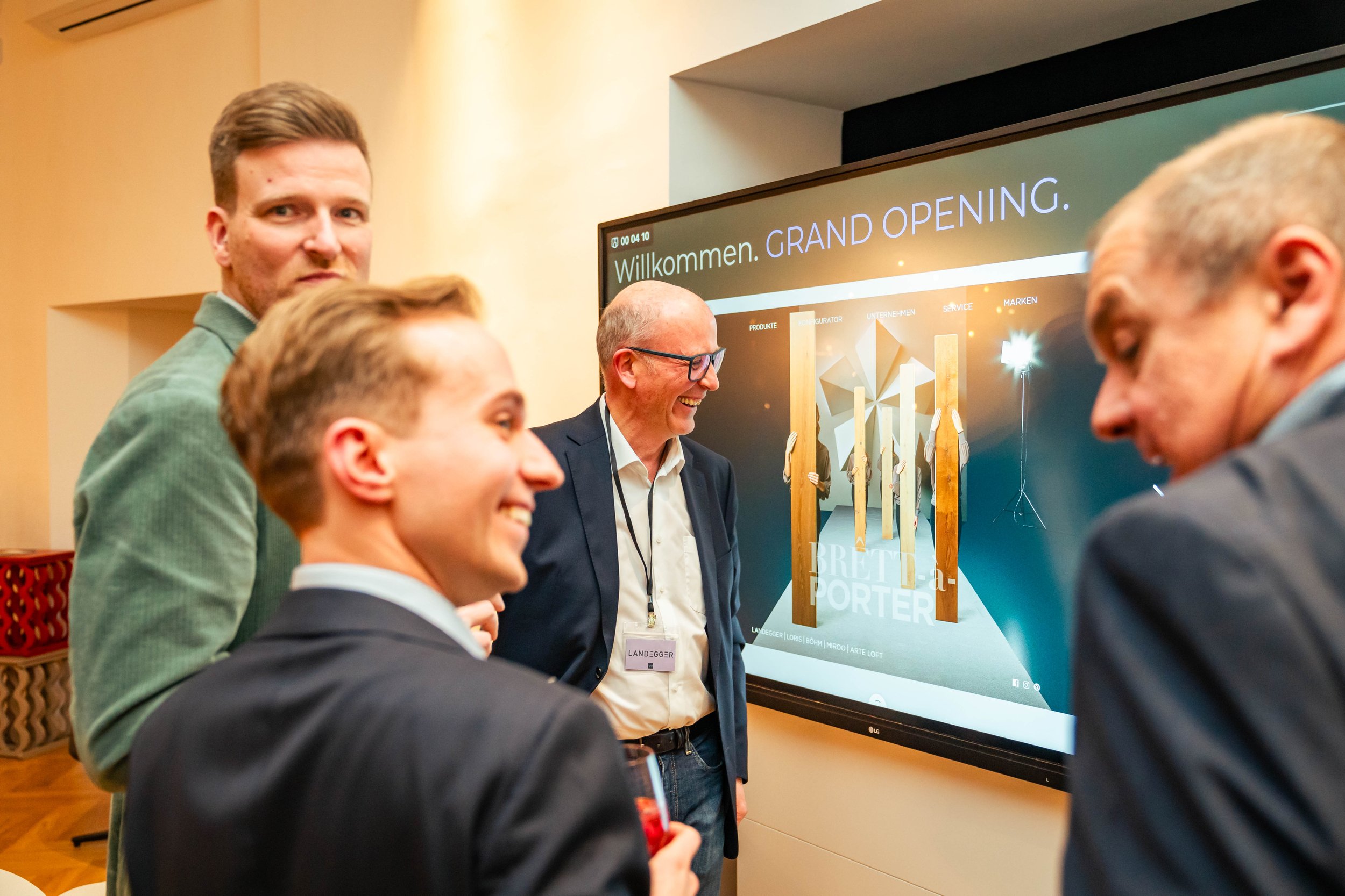 Vier Männer in Businesskleidung bei einer Veranstaltung mit einem großen Bildschirm im Hintergrund, der die Worte "Willkommen. GRAND OPENING." zeigt.